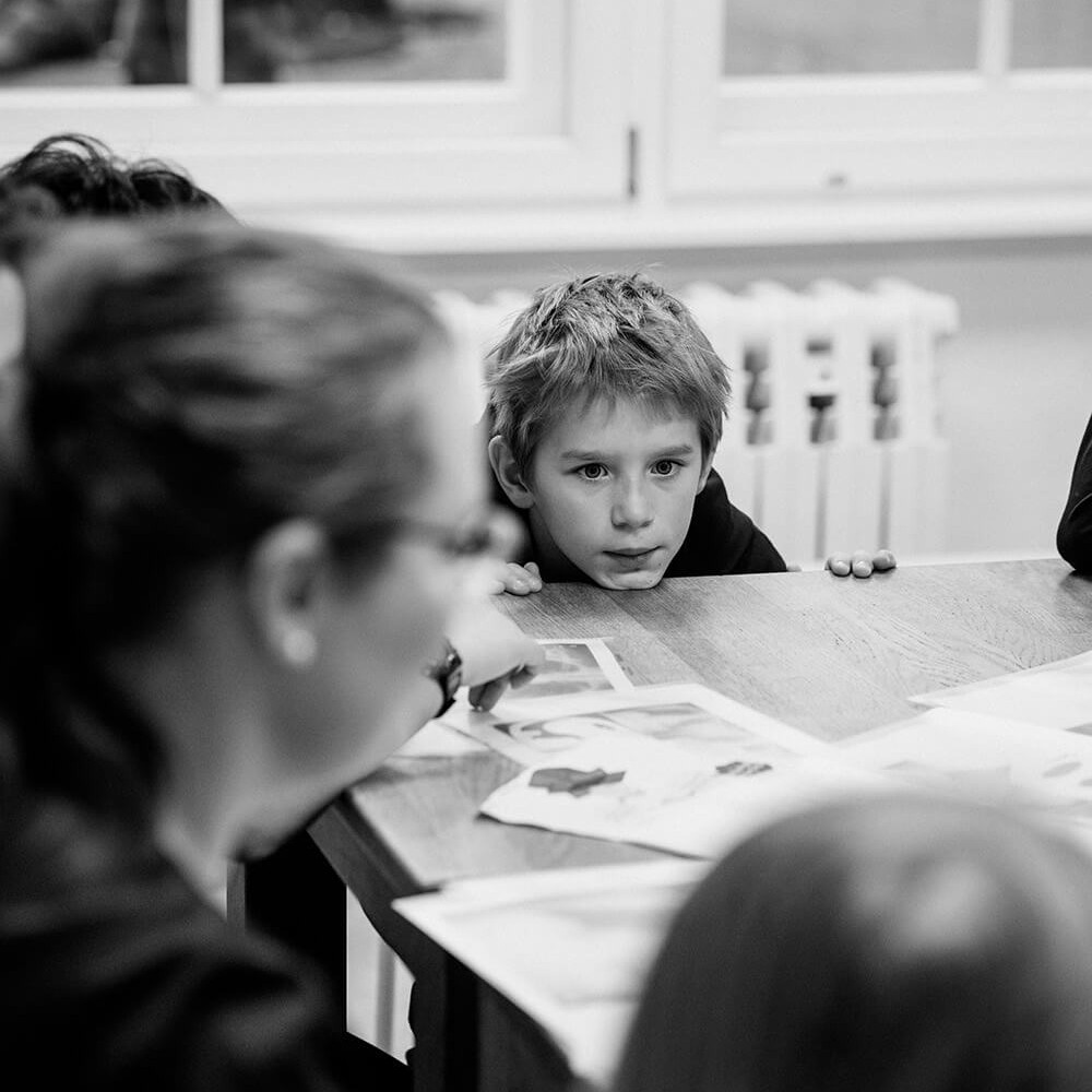 Cosmopolitanschool Berlin Ein Schwarz-Weiß-Foto eines Jungen, der auf dem BCS AI Day aufmerksam über einen mit Papieren bedeckten Tisch blickt, umgeben von anderen, die teilweise sichtbar sind, in einem Raum mit großen Fenstern, die auf die Zukunft dahinter hinweisen.