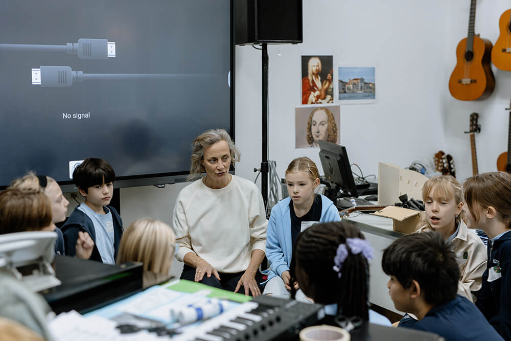 Cosmopolitanschool Berlin Ein Lehrer sitzt im Kreis mit aufmerksamen Schülern in einem BCS-Klassenzimmer, das mit Musikinstrumenten, Computern und einem großen Bildschirm mit der Meldung "Kein Signal" ausgestattet ist - eine moderne Umgebung für den KI-Tag und zukunftsorientiertes Lernen.