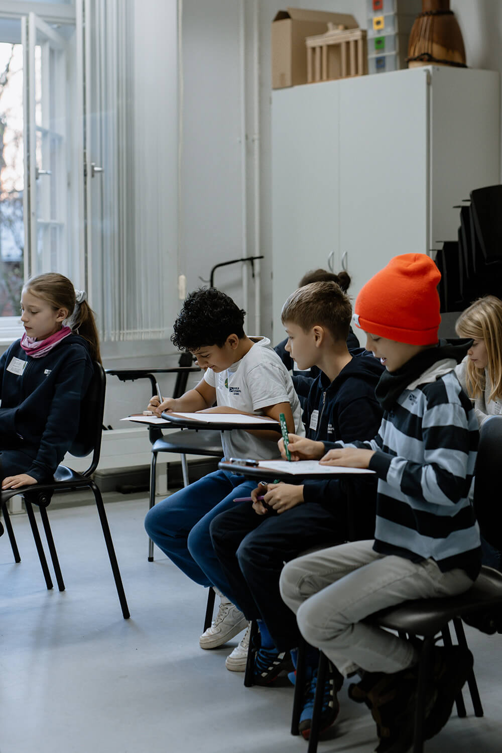 Cosmopolitanschool Berlin Eine Gruppe von Kindern sitzt in einem Klassenzimmer auf Stühlen mit angeschlossenen Tischen und schreibt oder zeichnet auf Papier. Ein Kind trägt eine orangefarbene Mütze. Im Hintergrund sind Regale und ein großes Fenster zu sehen, die während des BCS AI Day aufgestellt wurden.