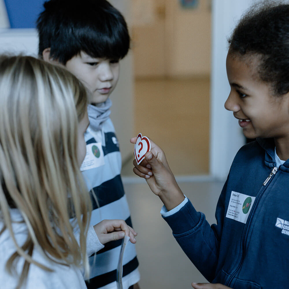 Cosmopolitanschool Berlin Drei Kinder stehen in einem Haus und unterhalten sich. Ein Mädchen lächelt und hält ein rundes Abzeichen mit einer roten "2" hoch, während die anderen zusehen. Alle drei tragen legere Kleidung und Namensschilder und unterhalten sich vielleicht über ihre Lieblingserlebnisse am AI Day bei BCS.
