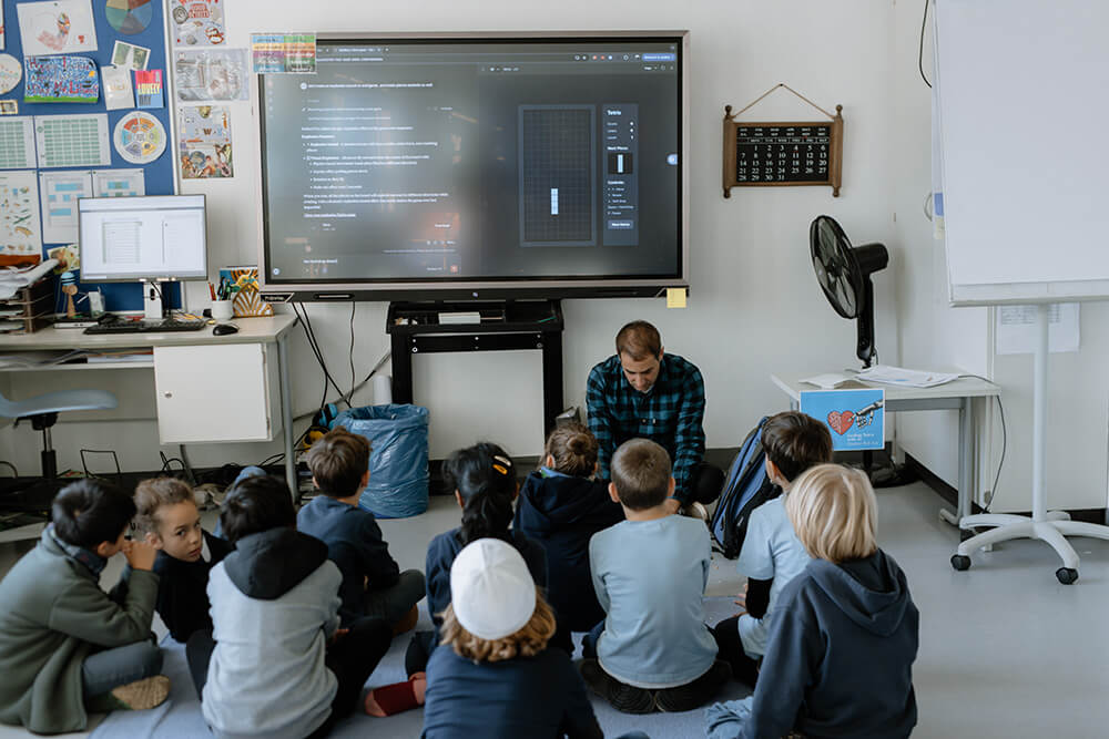 Cosmopolitanschool Berlin Ein Lehrer sitzt vor jungen Schülern auf dem Boden und blickt auf einen großen Bildschirm, auf dem während des KI-Tages in einem Klassenzimmer mit Computern, bunten Wanddekorationen und einem Kalender Code angezeigt wird - ein Blick in die Zukunft des Lernens.