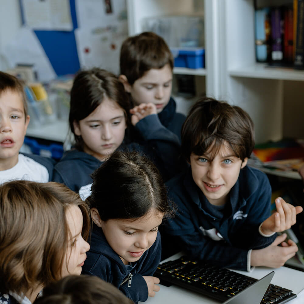 Cosmopolitanschool Berlin Eine Gruppe von Kindern in dunklen Kapuzenpullovern versammelt sich dicht um einen Laptop auf einem Schreibtisch. Sie wirken konzentriert und engagiert - einige lächeln und schauen auf den Bildschirm - während einer Veranstaltung zum KI-Tag, die von BCS in einem Klassenzimmer veranstaltet wurde, um gemeinsam die Zukunft zu erkunden. loading=