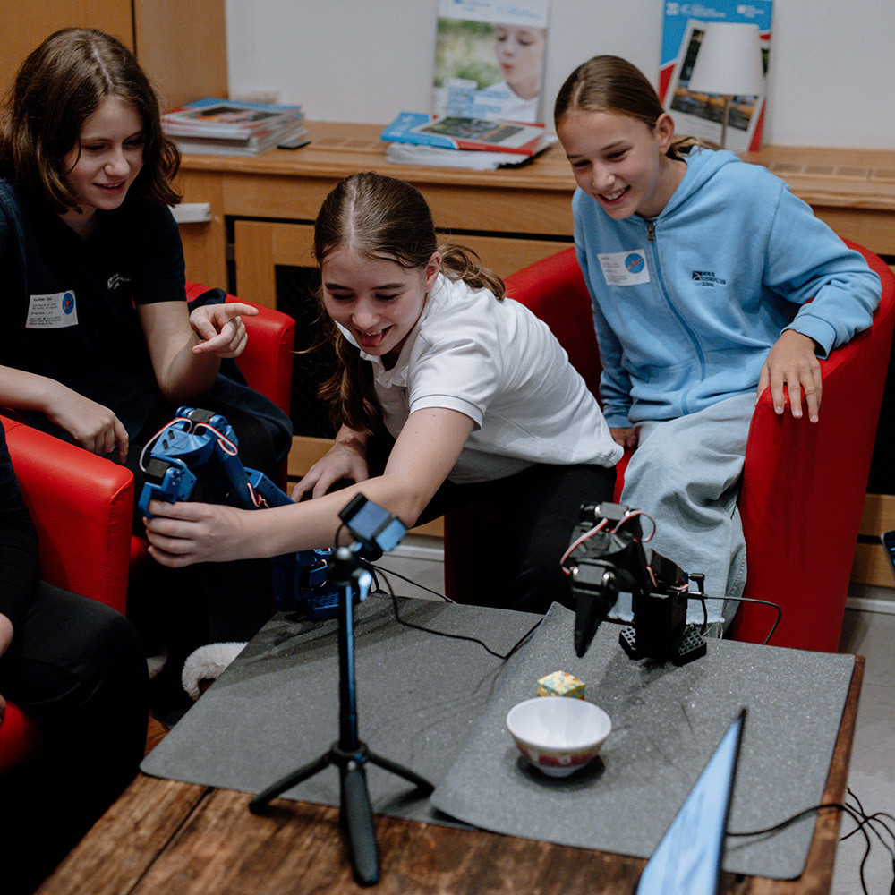 Cosmopolitanschool Berlin Drei Mädchen sitzen auf roten Stühlen, lächeln und interagieren mit einem kleinen Roboterarm auf einem Tisch während des AI Day. Ein Smartphone auf einem Stativ und ein Laptop stehen in der Nähe, im Hintergrund sind Bücher und Poster über künstliche Intelligenz zu sehen.