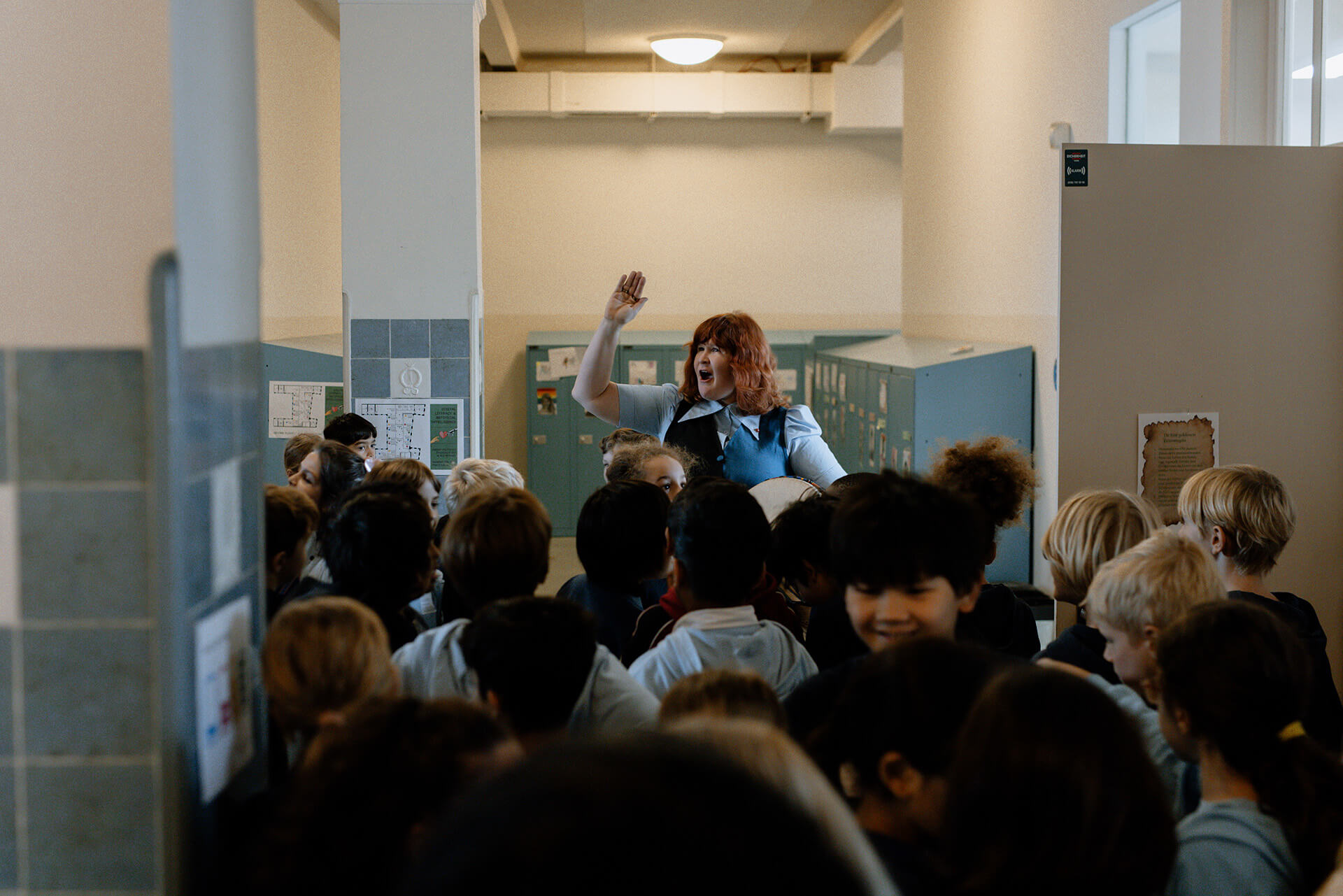 Cosmopolitanschool Berlin Eine Lehrerin steht in einem Schulflur, hebt eine Hand und spricht zu einer Gruppe von Kindern, die ihr gegenübersteht. Der Flur ist voller Schüler, und an den Wänden stehen Schließfächer. Die Atmosphäre wirkt lebendig und energiegeladen. loading=