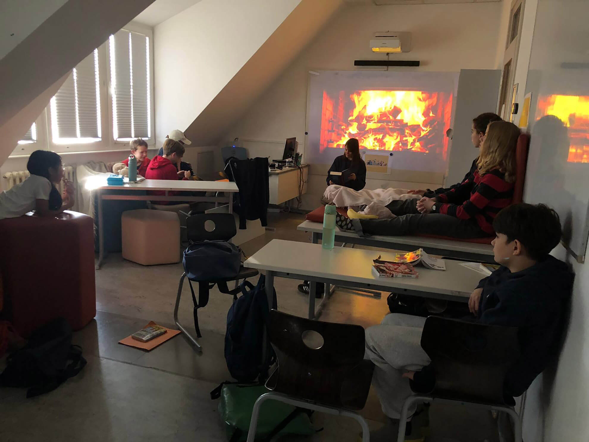 Cosmopolitanschool Berlin Eine Gruppe von Siebtklässlern entspannt sich in einem schwach beleuchteten Klassenzimmer. Einige sitzen an Tischen, andere lümmeln auf Sofas. Ein digitaler Kamin und verstreute Lieblingsbücher schaffen eine gemütliche Atmosphäre für den Vorlesetag. Taschen und Bücher liegen verstreut herum.