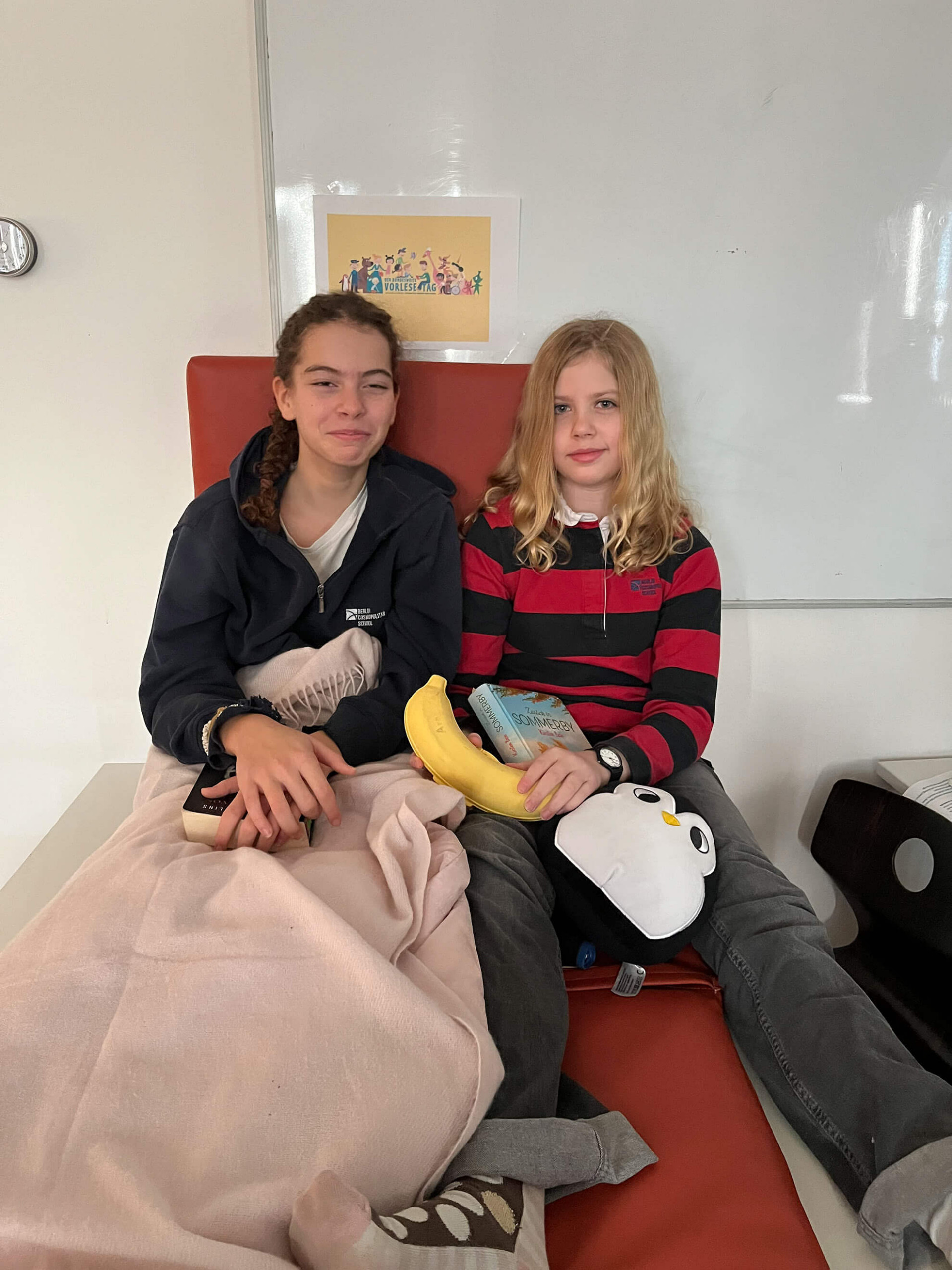 Cosmopolitanschool Berlin Zwei Kinder sitzen nebeneinander auf einem orangefarbenen Stuhl. Es ist Vorlesetag, und beide lächeln leicht - das eine hält seine Lieblingsbücher und eine Decke in der Hand, das andere hat Bananen, ein Buch und einen Pinguin aus Plüsch. Hinter ihnen steht eine Tafel mit buntem Druck.
