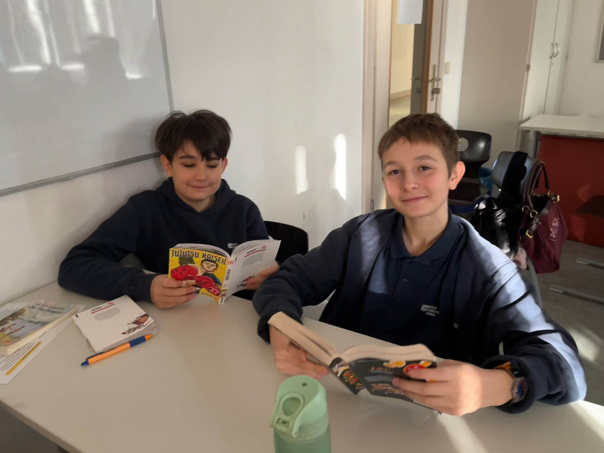 Cosmopolitanschool Berlin Zwei Siebtklässler sitzen an einem Klassenzimmerpult, lächeln in die Kamera und halten ihre Lieblingsbücher in der Hand. Einer trägt einen marineblauen Kapuzenpulli, der andere eine blaue Jacke. Auf dem Pult liegen Schulsachen und eine grüne Wasserflasche, während von links das Sonnenlicht hereinstrahlt.