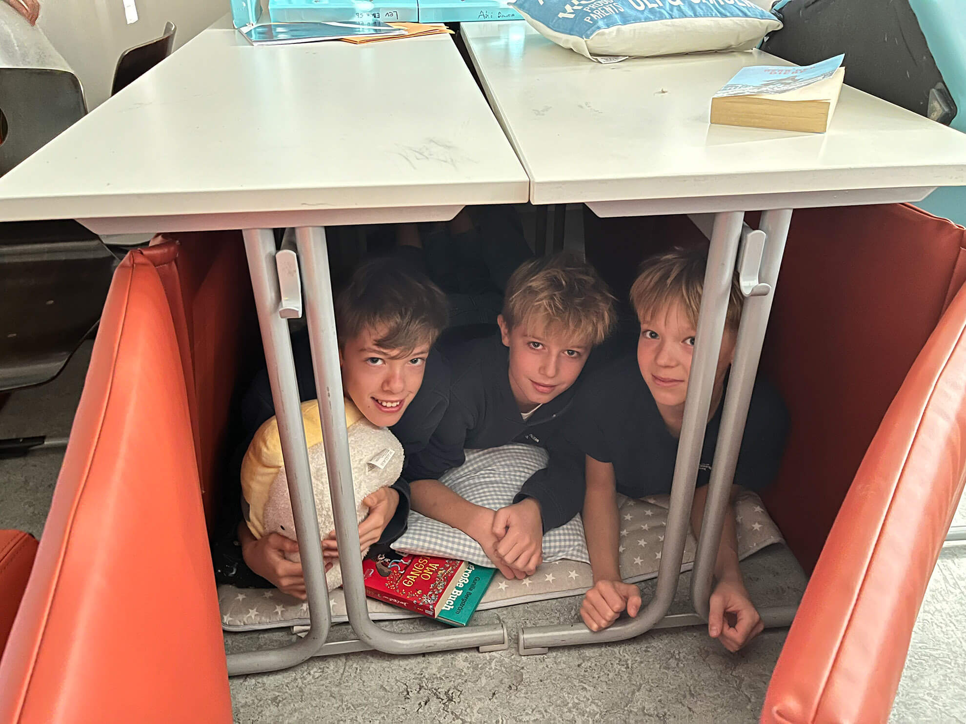 Cosmopolitanschool Berlin Drei Siebtklässler sitzen eng beieinander unter einem Schreibtisch, umgeben von orangefarbenen Kissen, mit Kissen und ihren Lieblingsbüchern. Sie haben einen gemütlichen, festungsartigen Raum geschaffen und alle drei schauen in die Kamera und lächeln.