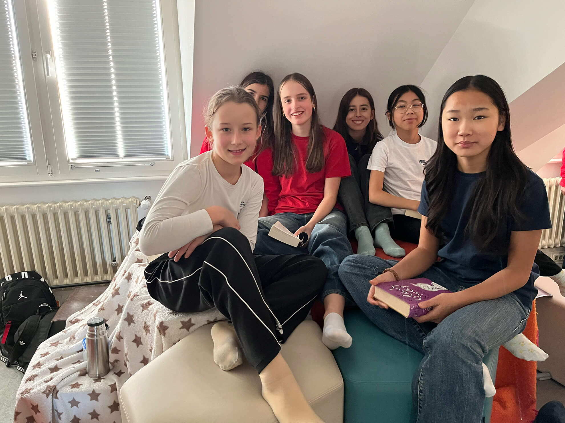 Cosmopolitanschool Berlin Sechs Siebtklässler sitzen eng beieinander auf einer gemütlichen, gut beleuchteten Couch, einige halten ihre Lieblingsbücher und lächeln in die Kamera. Ein Teil der Couch ist mit einer Sternendecke bedeckt, daneben stehen Taschen und eine Wasserflasche auf dem Boden.