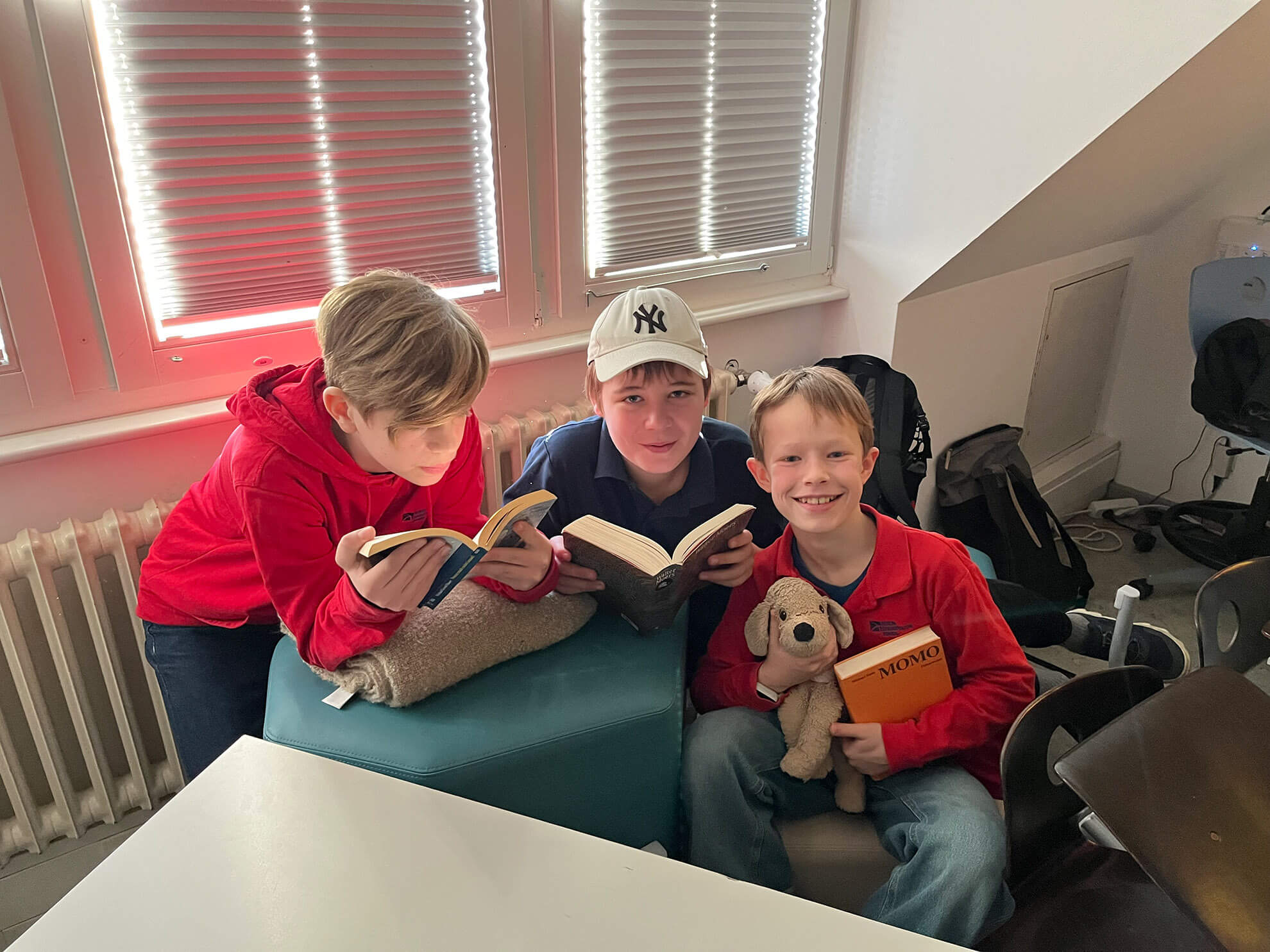Cosmopolitanschool Berlin Drei Jungen sitzen drinnen an einem Fenster und lesen Bücher, als ob sie ihre eigenen kleinen Höhlen schaffen würden. Der Junge in der Mitte trägt eine weiße Mütze, während der Junge rechts einen braunen Stoffhund und ein Buch mit dem Titel "Atomoe" in der Hand hält - wie geschaffen für einen gemütlichen Vorlesetag.