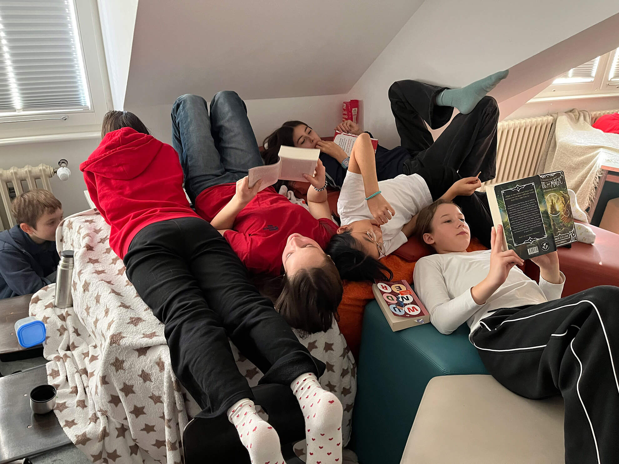 Cosmopolitanschool Berlin Fünf Siebtklässler sitzen eng beieinander auf einem Sofa und Kissen in einem gemütlichen, sonnendurchfluteten Raum, jeder vertieft in die Lektüre seiner Lieblingsbücher. Sie wirken entspannt und bequem, genießen einen gemeinsamen Moment der Ruhe und Konzentration. loading=