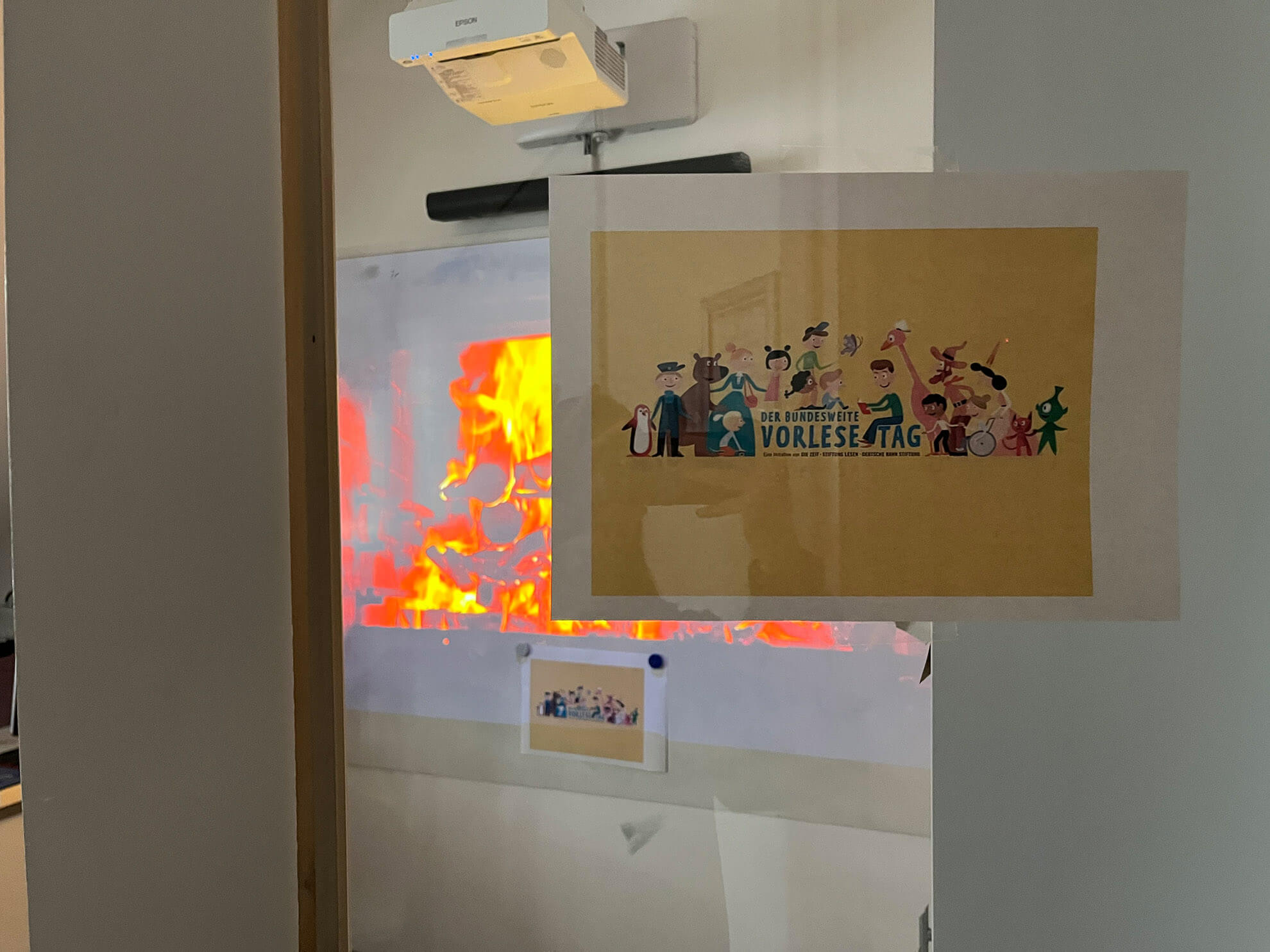 Cosmopolitanschool Berlin Eine Tür mit einem Plakat mit Zeichentrickfiguren und dem deutschen Text "Vorlesetag" lädt Siebtklässler ein, ihre Lieblingsbücher mitzuteilen. Durch eine Glasscheibe ist im Inneren des Raumes ein Bildschirm mit animierten Flammen zu sehen.