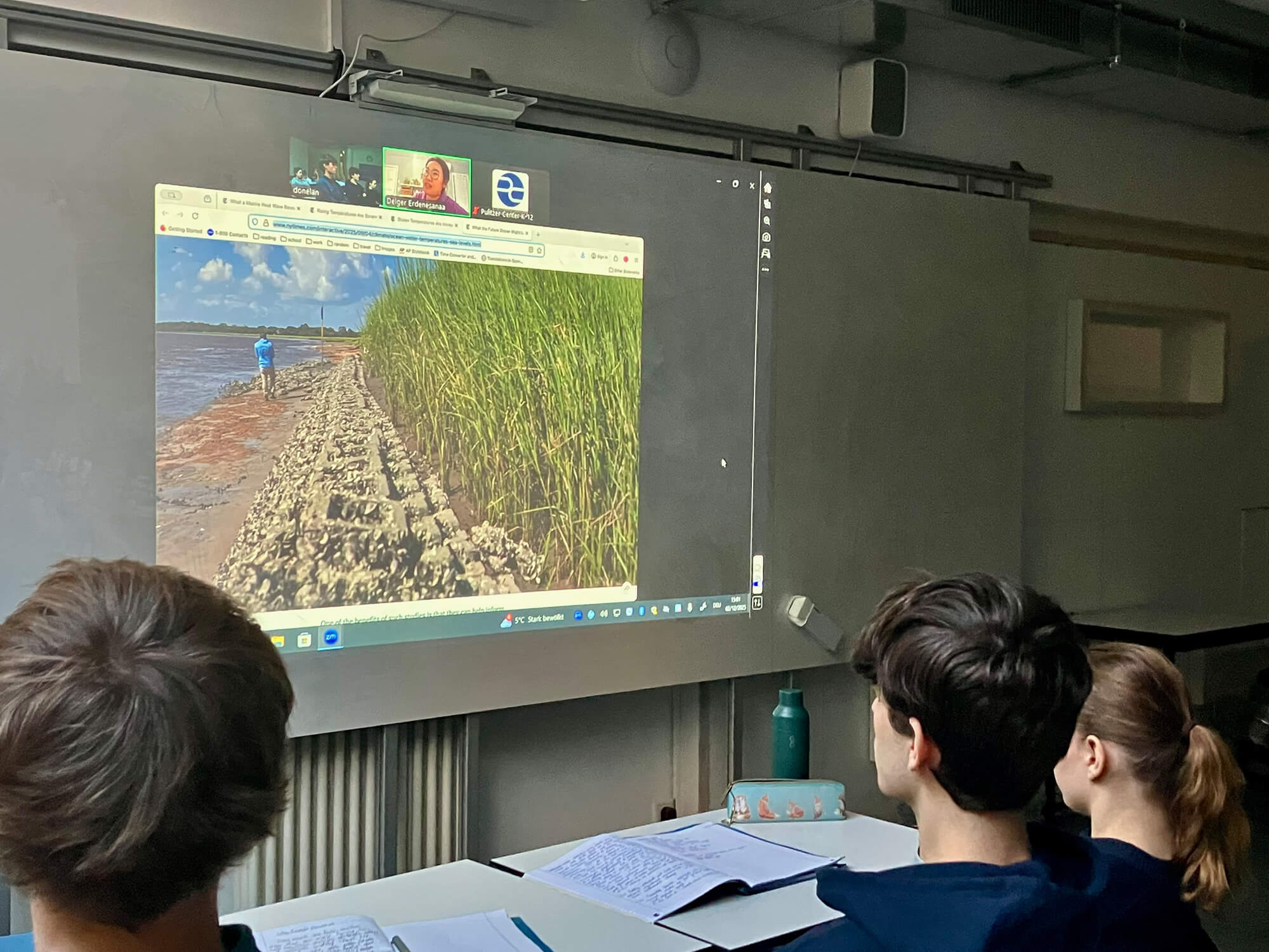 Cosmopolitanschool Berlin Drei Schüler einer BCS-Abiturklasse sitzen an ihren Tischen und sehen sich ein projiziertes Video auf einem Whiteboard an. Ein Reporter der "New York Times" ist zu sehen, wie er einen felsigen Weg entlang von Wasser und hohem Gras entlangläuft, wobei die Gesichter zweier Personen in kleinen Videofenstern zu sehen sind. mit KI bearbeitet