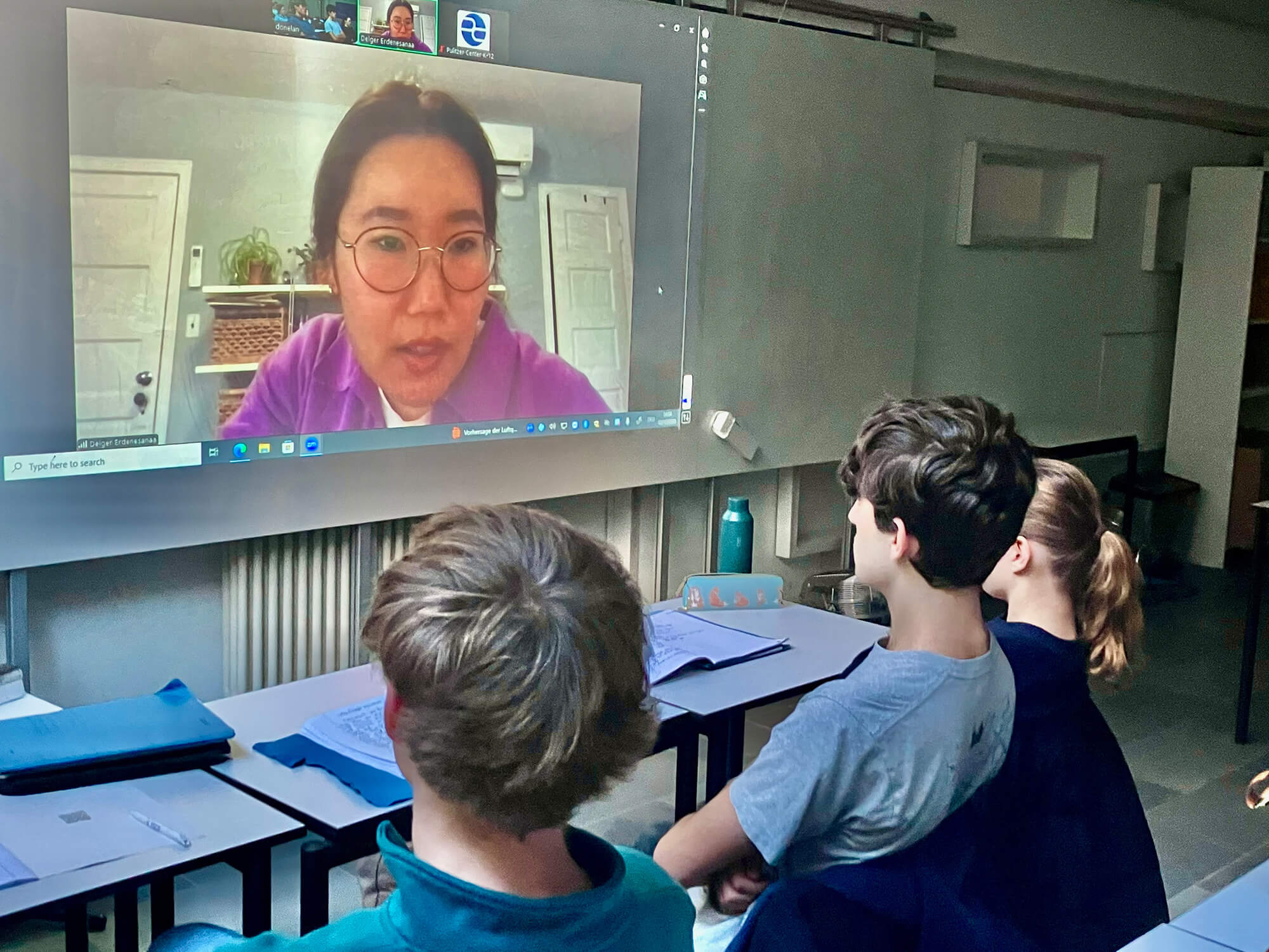Cosmopolitanschool Berlin Drei Schüler einer BCS-Abiturklasse sitzen an ihren Tischen und sehen sich ein projiziertes Video auf einem Whiteboard an. Ein Reporter der "New York Times" ist zu sehen, wie er einen felsigen Weg entlang von Wasser und hohem Gras entlangläuft, wobei die Gesichter zweier Personen in kleinen Videofenstern zu sehen sind. mit KI bearbeitet