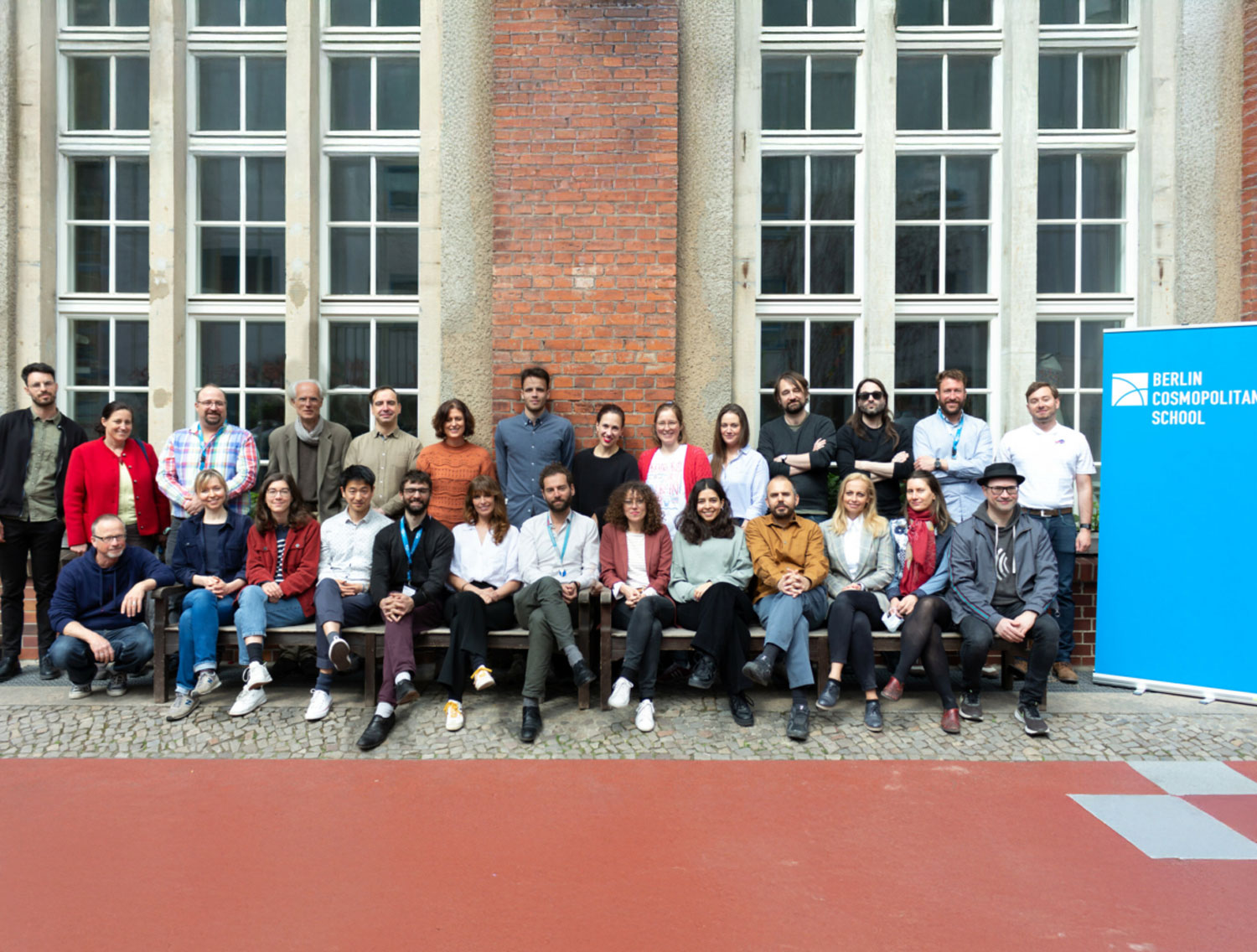 Cosmopolitanschool Berlin Eine Gruppe von etwa 30 Erwachsenen posiert gemeinsam vor einem Backsteingebäude mit hohen Fenstern. Sie sitzen und stehen in Reihen. Auf einem blauen Schild auf der rechten Seite steht "Berlin Cosmopolitan School".