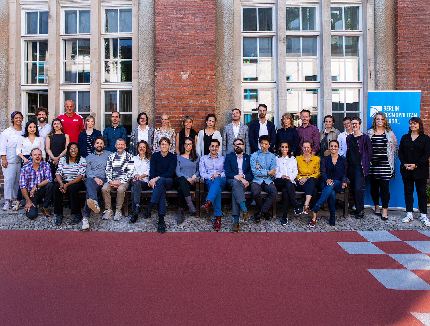 Cosmopolitanschool Berlin Eine Gruppe von etwa 35 Erwachsenen posiert für ein Foto vor einem Backsteingebäude mit großen Fenstern. Einige Personen sitzen, während andere hinter ihnen stehen. Auf einem blauen Transparent steht "Berlin Cosmopolitan School".