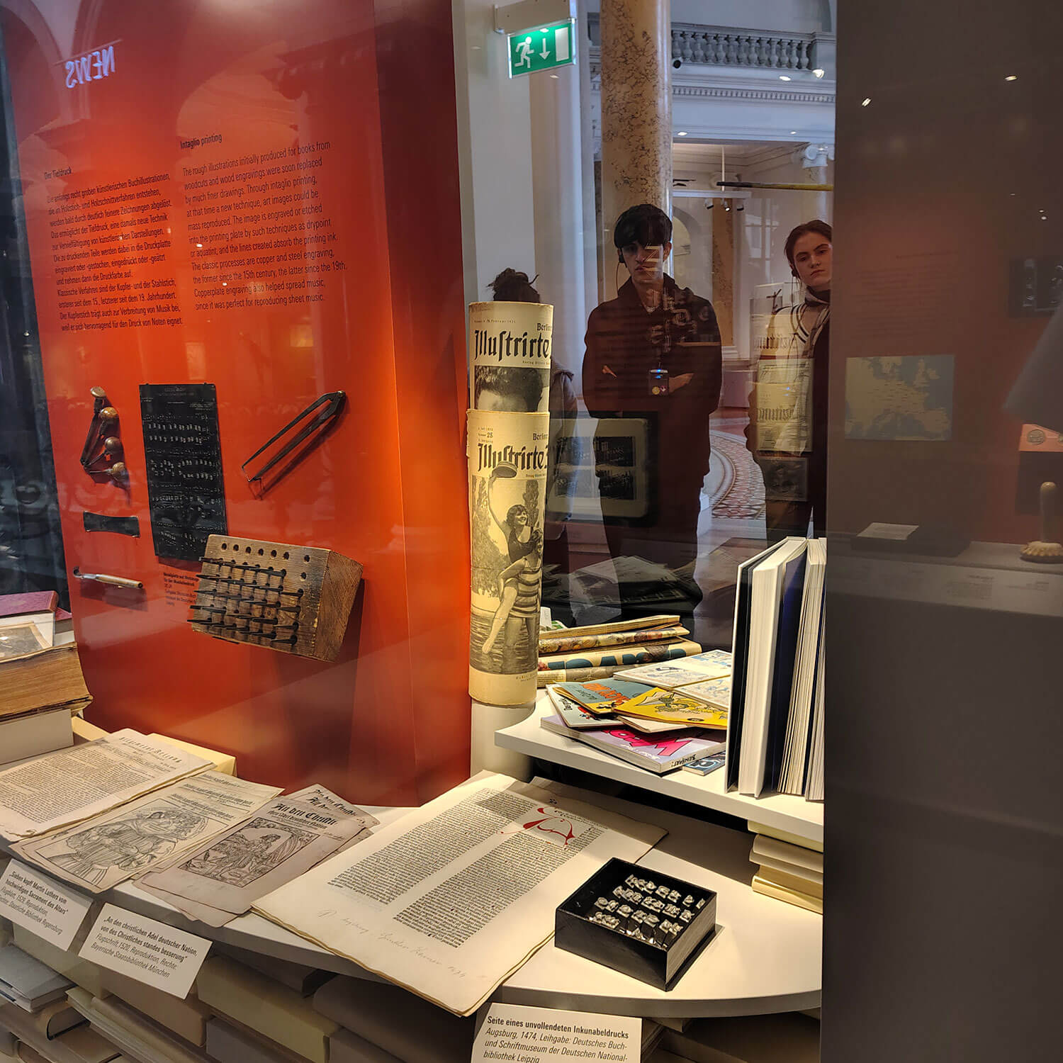 Cosmopolitanschool Berlin Eine Museumsvitrine mit alten Büchern, Zeitungen und Artefakten - Medieninstrumente der Macht - zeigt eine Schreibmaschine und einen Nummernstempel. Zwei Personen spiegeln sich im Glas und betrachten das Exponat "World of Media" vor einer orangefarbenen Informationstafel.