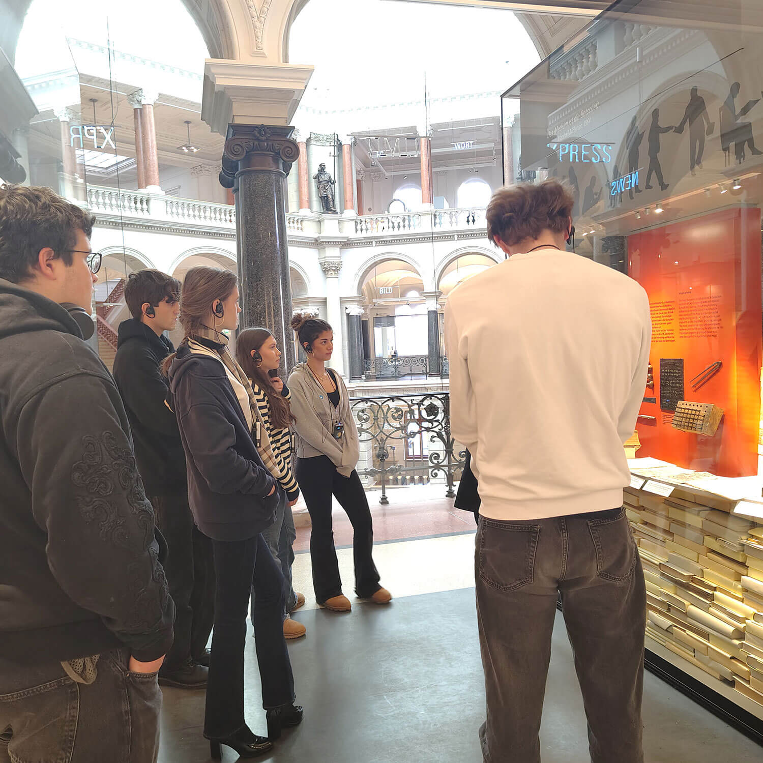 Cosmopolitanschool Berlin In einem geräumigen, reich verzierten Museum mit hohen Säulen und gewölbten Balkonen steht eine Gruppe von Menschen und betrachtet eine Ausstellung über Medieninstrumente der Macht. Die Vitrine enthält historische Artefakte und hell erleuchtete Informationsdisplays.