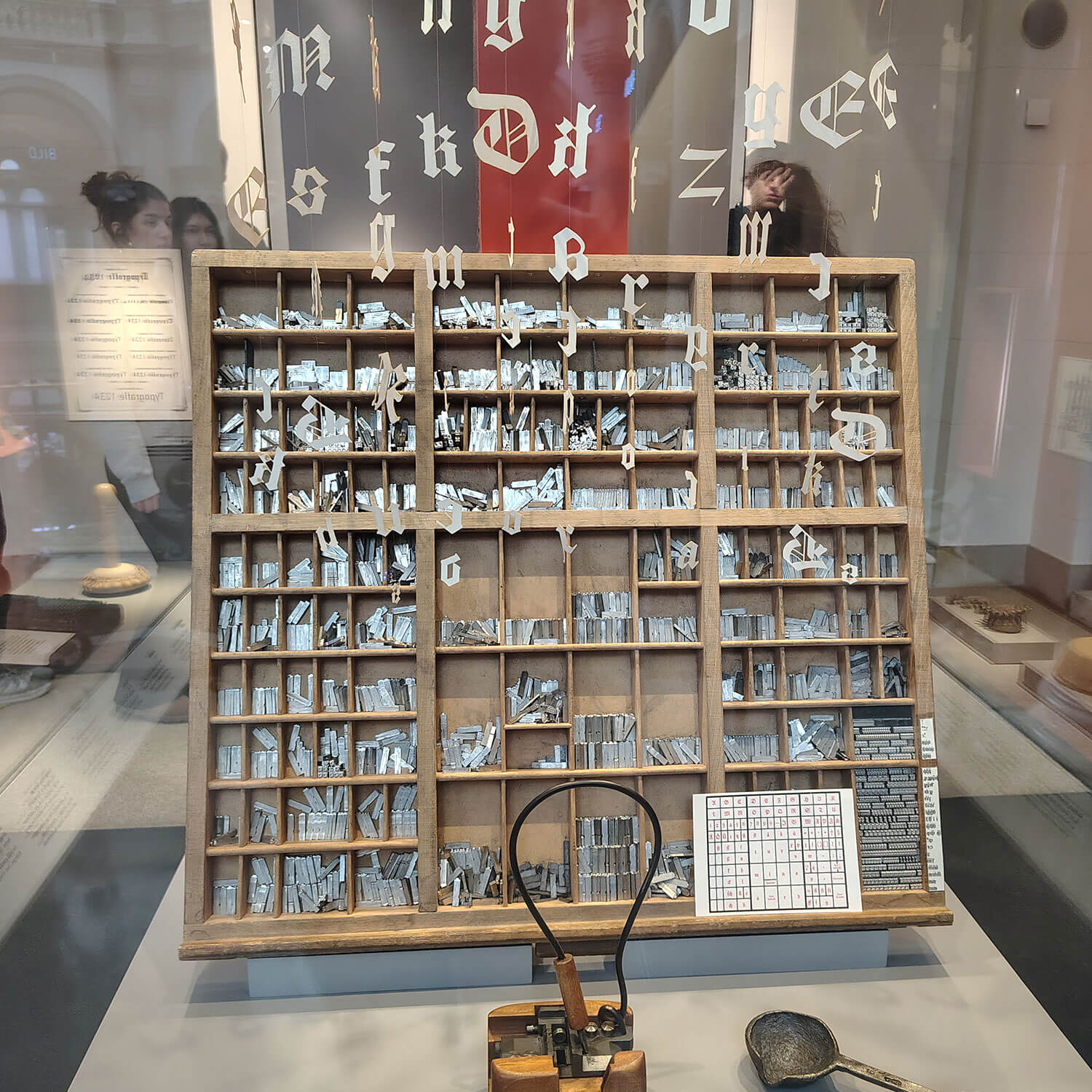 Cosmopolitanschool Berlin Eine Holzkiste mit beweglichen Metalltypen für den Buchdruck, die im Rahmen der Ausstellung World of Media in einem Museum ausgestellt ist, mit Buchstaben im gotischen Stil auf dem Glas dahinter und Druckwerkzeugen im Vordergrund.