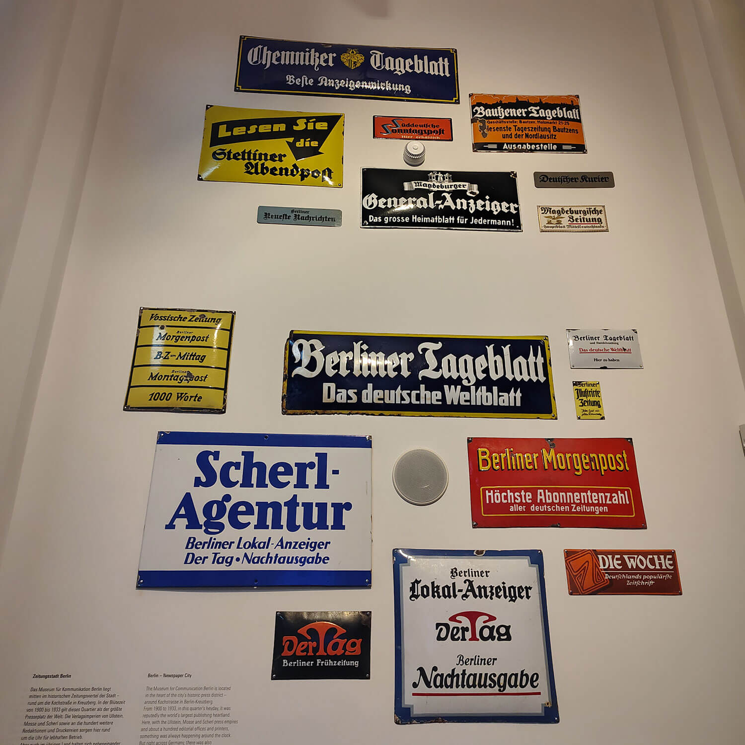 Cosmopolitanschool Berlin Eine Ausstellung alter deutscher Zeitungs- und Werbeschilder in verschiedenen Größen, Formen und Schriftarten, die an einer weißen Wand angebracht sind. Das Exponat hebt mediale Machtinstrumente mit Namen wie Berliner Tageblatt und Scherl Agentur hervor.
