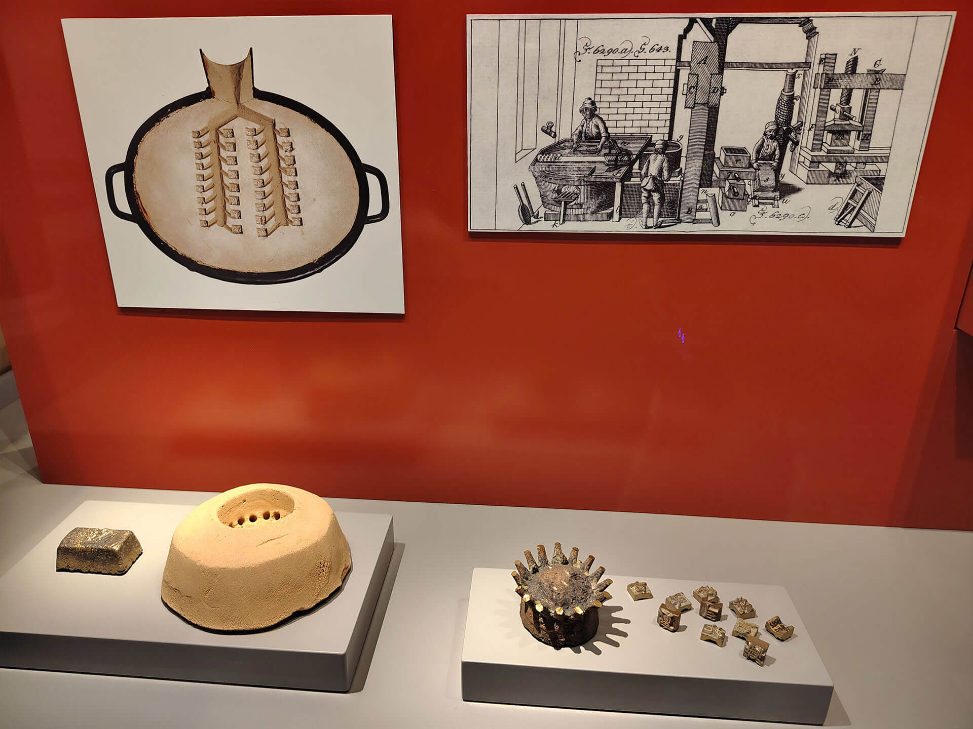 Cosmopolitanschool Berlin Eine Museumsausstellung zeigt historische Metallformen, Gold- oder Silberbarren und einen Abdruck einer Form unter einer alten Schwarz-Weiß-Illustration der Metallverarbeitung auf einer roten Wand, die die Welt der Medien als Instrumente der Macht im Laufe der Geschichte hervorhebt.