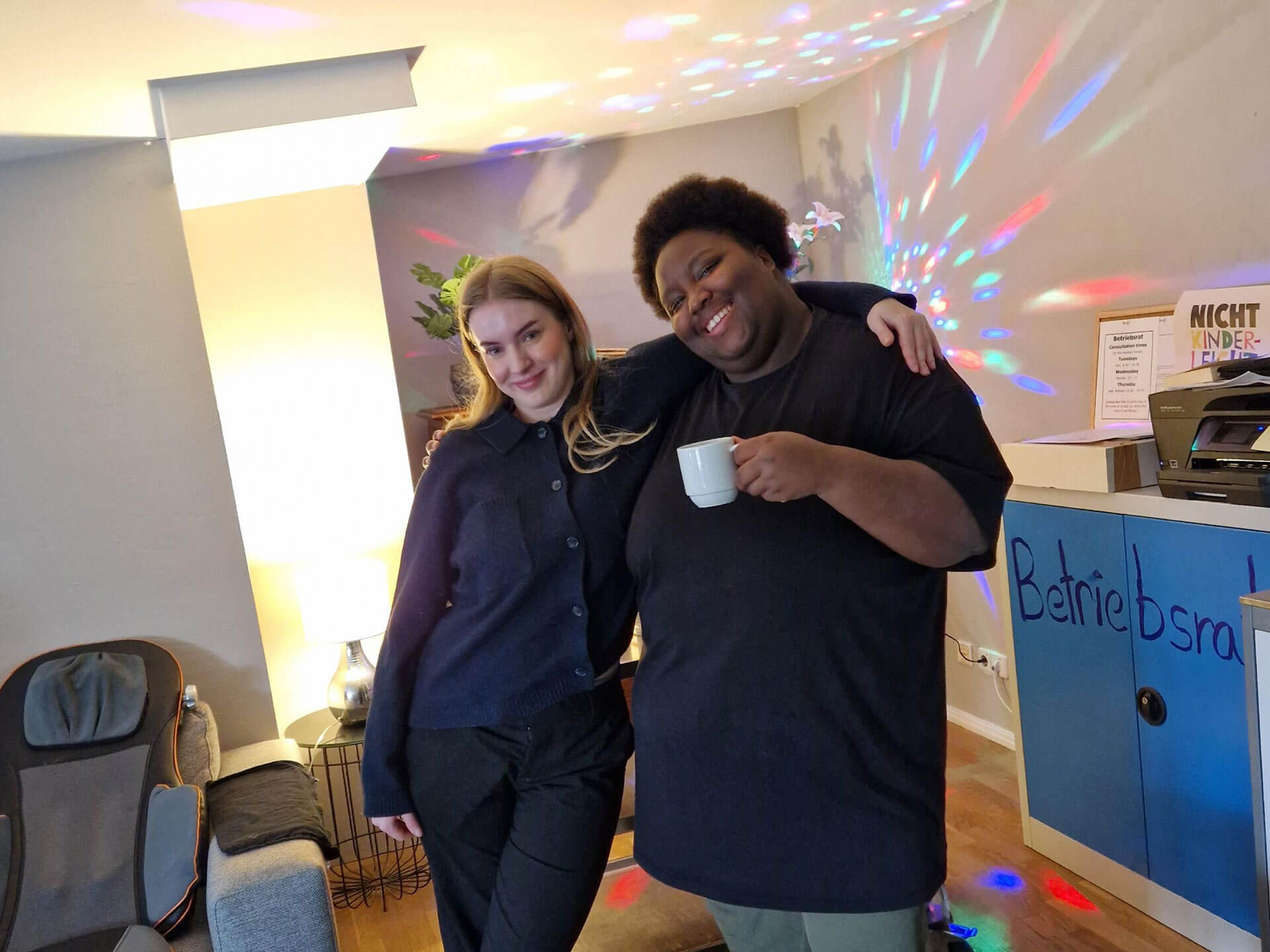 Cosmopolitanschool Berlin Zwei Menschen stehen dicht beieinander und lächeln während der BCS Kita-Auszubildenden Weihnachtsfeier in die Kamera. Einer hält einen weißen Becher, während hinter ihnen bunte Lichter an der Wand tanzen. In dem gemütlichen Raum sind ein blauer Tresen und ein Stuhl zu sehen.