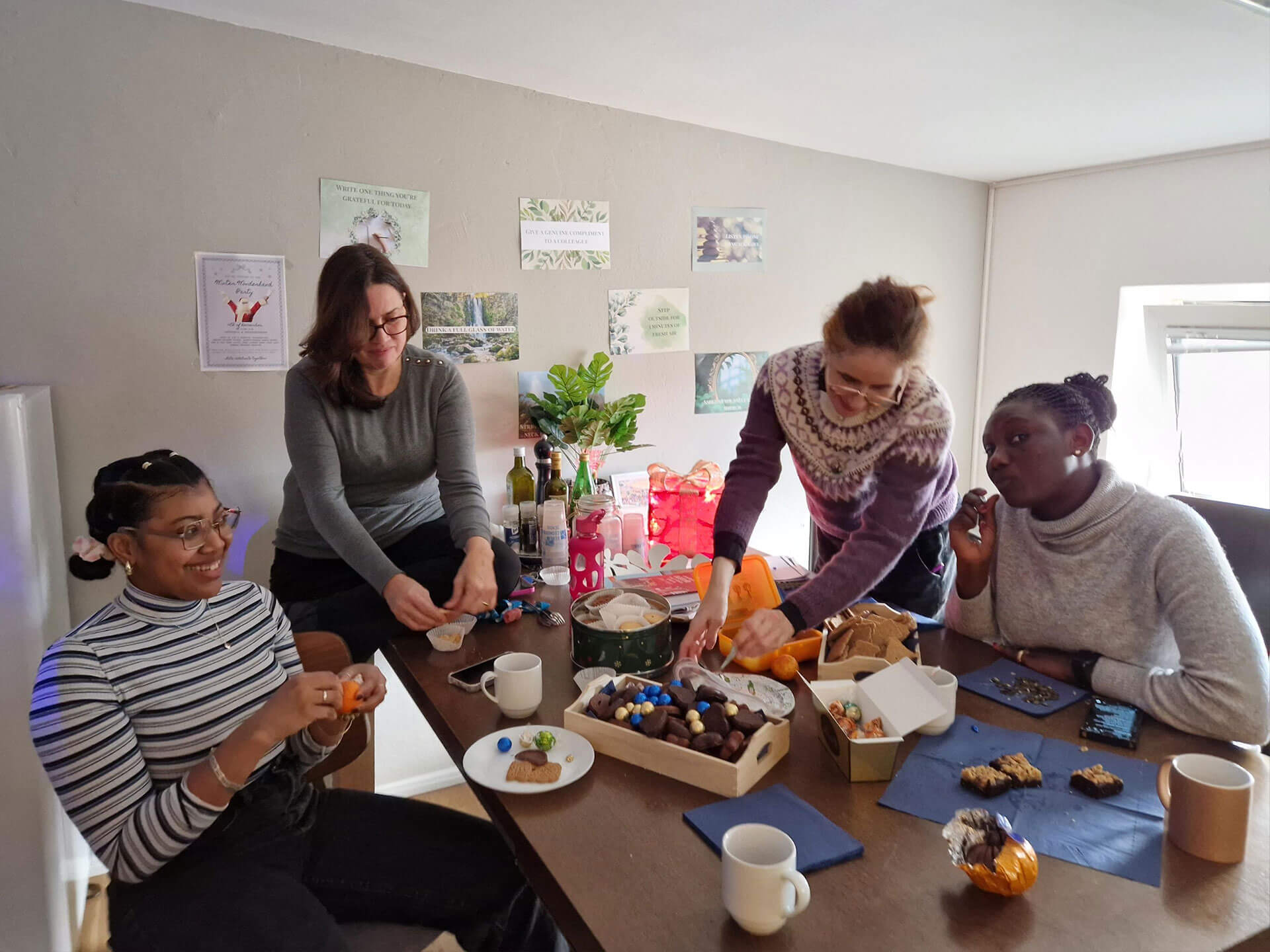 Cosmopolitanschool Berlin Vier Frauen sitzen und stehen bei der BCS Kita-Auszubildenden Weihnachtsfeier um einen mit Snacks, Gebäck und Getränken gedeckten Tisch, plaudern und bedienen sich in einem gemütlichen, hellen, mit kleinen Postern und Pflanzen dekorierten Raum.