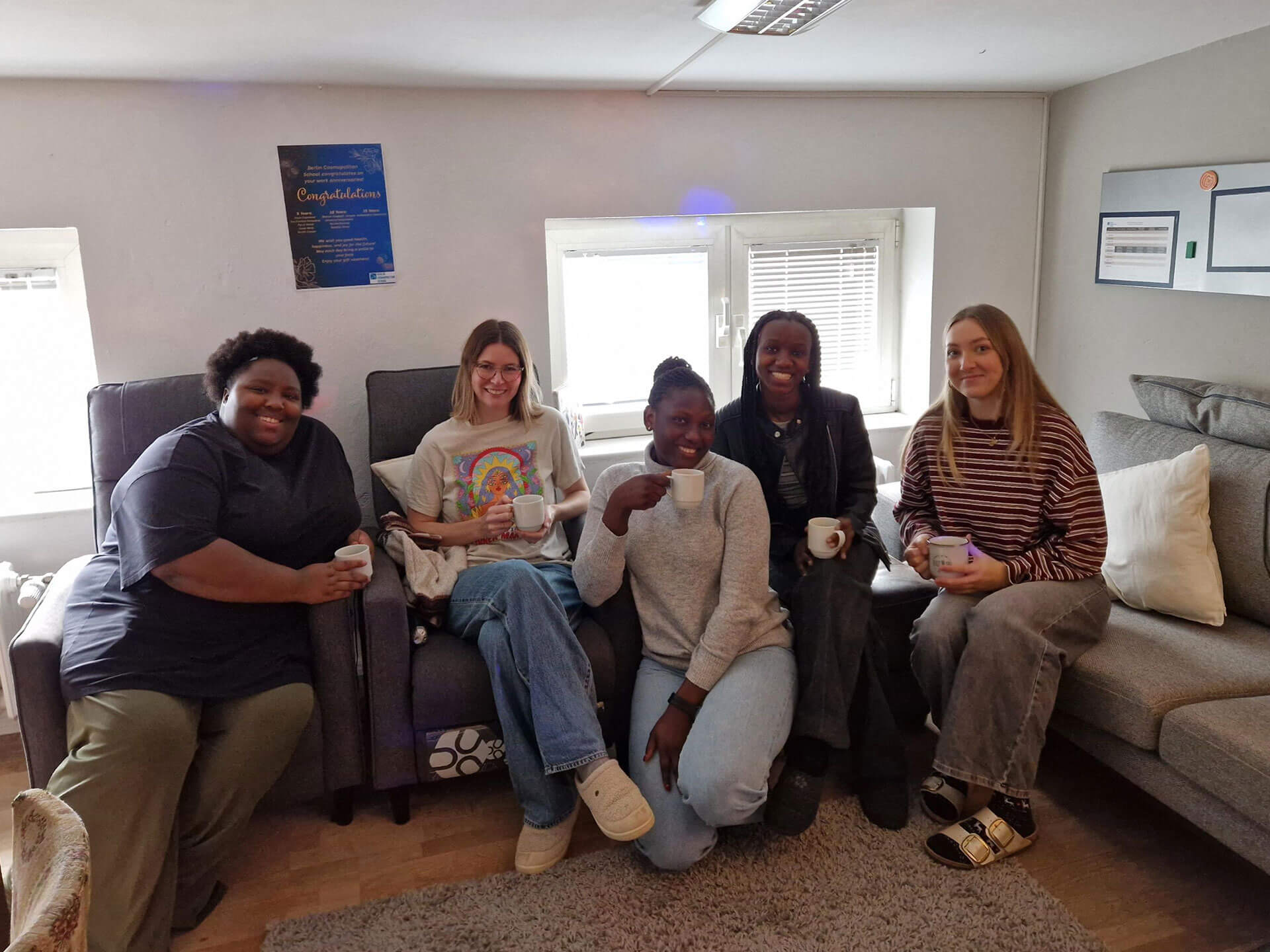 Cosmopolitanschool Berlin Fünf Frauen, wahrscheinlich BCS Kita-Auszubildende, sitzen auf Sofas in einem gemütlichen, gut beleuchteten Raum während eines Jahresabschlusses. Sie lächeln und halten Tassen in der Hand, hinter ihnen hängen Urkunden und Poster an der Wand.