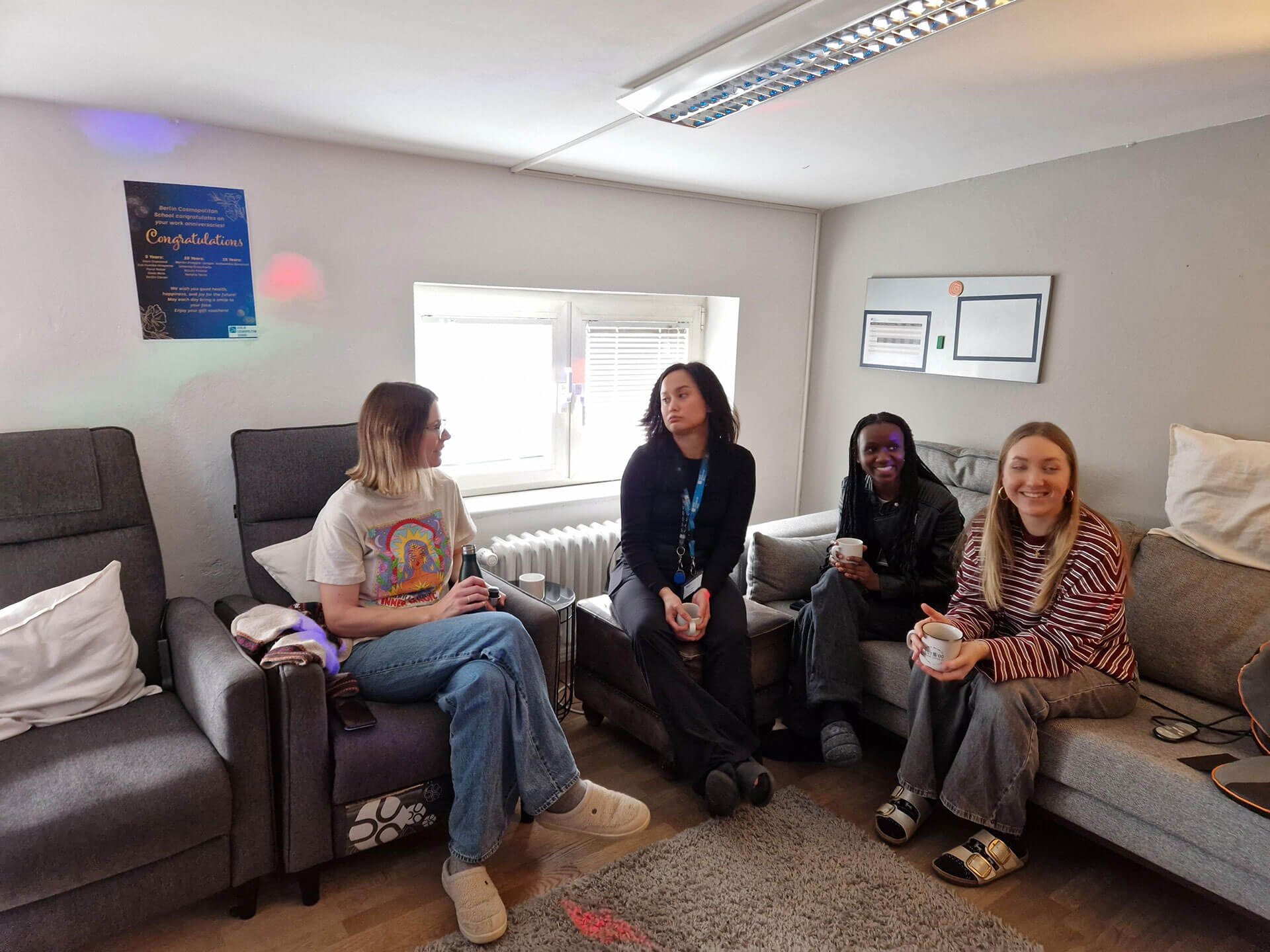 Cosmopolitanschool Berlin Vier Frauen sitzen auf Sofas in einem gemütlichen Raum mit grauen Wänden, genießen Tassen und lächeln während des BCS Kita-Auszubildenden Jahresabschlusses. Natürliches Licht strömt durch das Fenster, an der Wand hängen ein Poster und Urkunden.