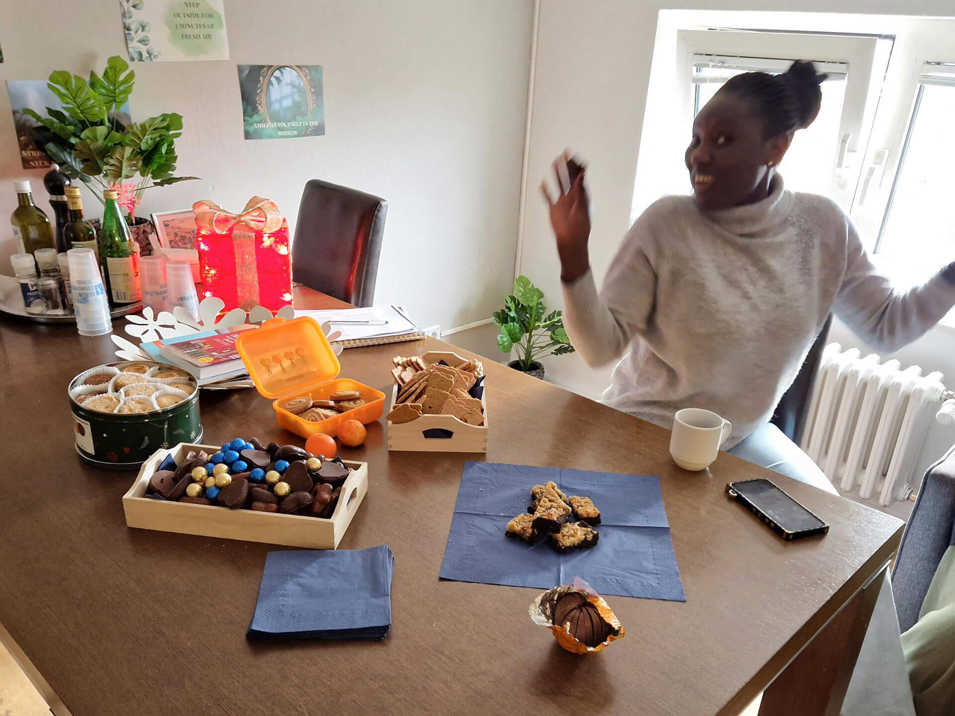 Cosmopolitanschool Berlin Eine Frau lächelt und gestikuliert energisch, während sie an einem Tisch mit Keksen, Schokolade, Obst, einem Kaffeebecher und einem Smartphone sitzt - und damit die festliche Stimmung der BCS Kita-Auszubildenden Jahresabschluss Weihnachtsfeier einfängt. Pflanzen und Dekorationen erhellen den Raum. loading=