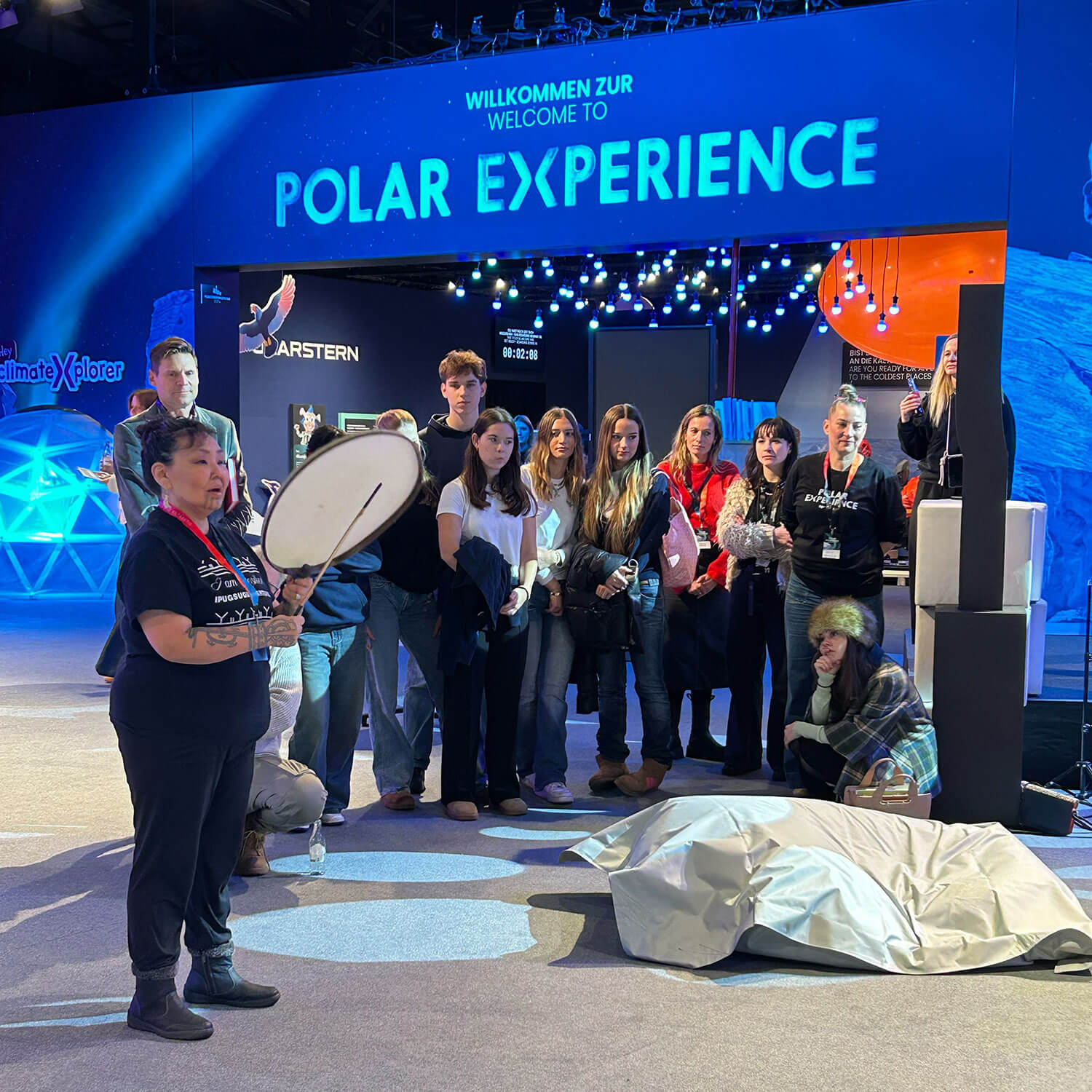 Cosmopolitanschool Berlin Eine Gruppe von Menschen hört einem Vortragenden zu, der eine Handtrommel vor dem Polar Experience Exponat hält, während blaue Beleuchtung und ein Willkommensschild die Szene einrahmen - ein automatisch gespeicherter Entwurf für inspirierende Veranstaltungen. loading=