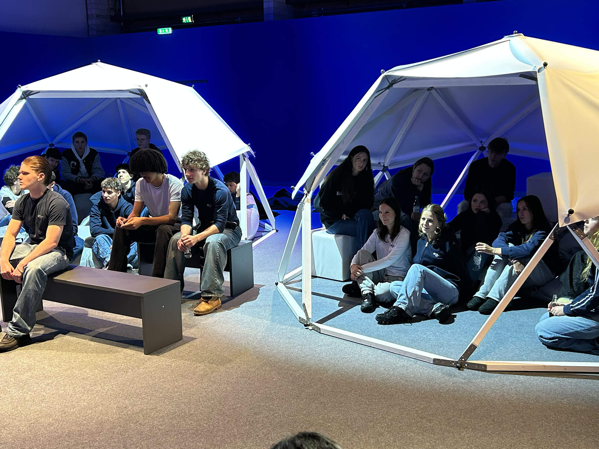 Cosmopolitanschool Berlin Eine Gruppe junger Menschen sitzt auf Bänken und in geodätischen Kuppelstrukturen unter hellen Lichtern. Sie wirken aufmerksam und engagiert in einem modernen Innenraum mit blauem Hintergrund - wie ein automatisch gespeicherter Entwurf, der zum Leben erweckt wurde. loading=