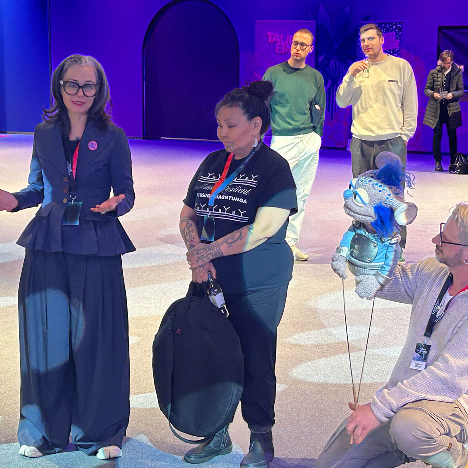 Cosmopolitanschool Berlin Eine Frau in einem marineblauen Anzug spricht mit Gesten zu einer Gruppe in einem Haus. Neben ihr hält eine Frau in einem schwarzen Hemd eine Tasche mit der Aufschrift "Automatisch gespeicherter Entwurf", während ein Mann mit einer blauen Puppe in der Hand kniet. Drei Personen stehen unter violettem Licht.