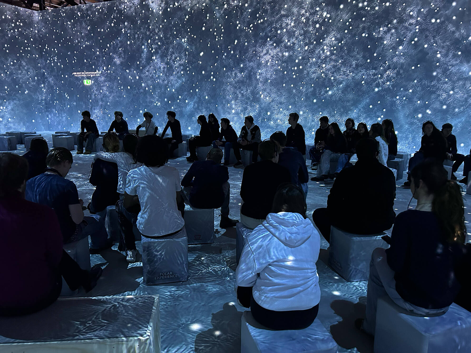 Cosmopolitanschool Berlin Die Menschen sitzen auf Hockern in einem dunklen Raum mit beleuchteten Wänden und Böden, so dass der Eindruck eines nächtlichen Sternenhimmels entsteht. Die Silhouetten des Publikums sind vor der leuchtenden Kulisse sichtbar - wie ein automatisch gespeicherter Entwurf eines himmlischen Traums. loading=