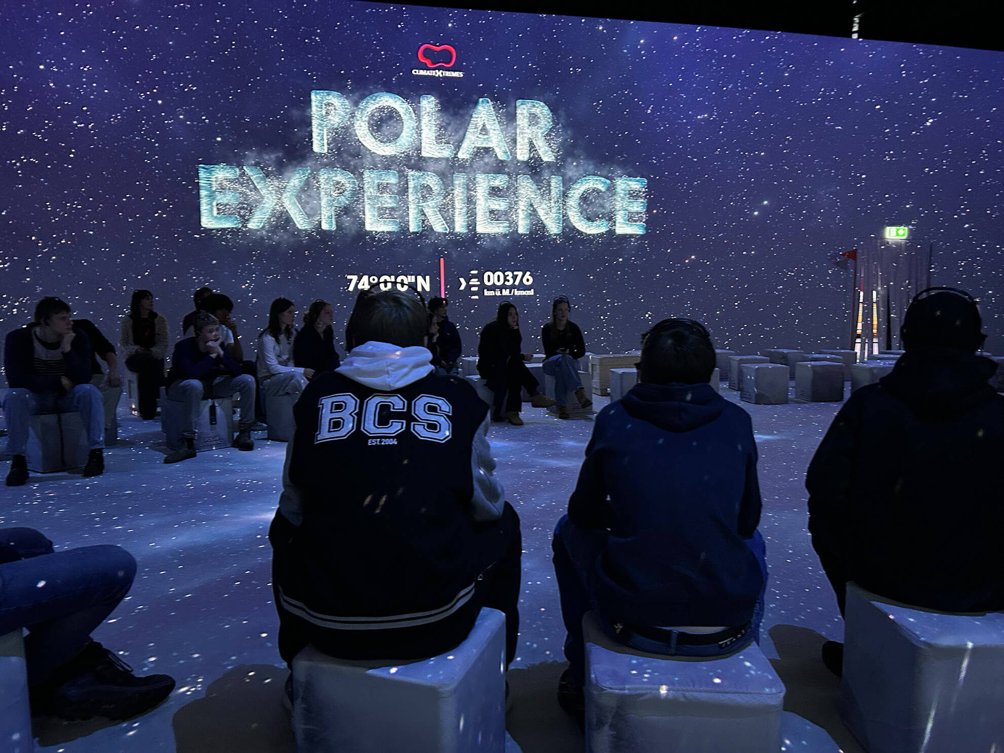 Cosmopolitanschool Berlin Menschen sitzen auf weißen Hockern in einem dunklen Raum mit schneeähnlichen Lichtprojektionen. POLAR EXPERIENCE wird in großen Buchstaben neben einem automatisch gespeicherten Entwurf angezeigt und schafft so eine immersive und winterliche Atmosphäre mit Koordinaten darunter. loading=