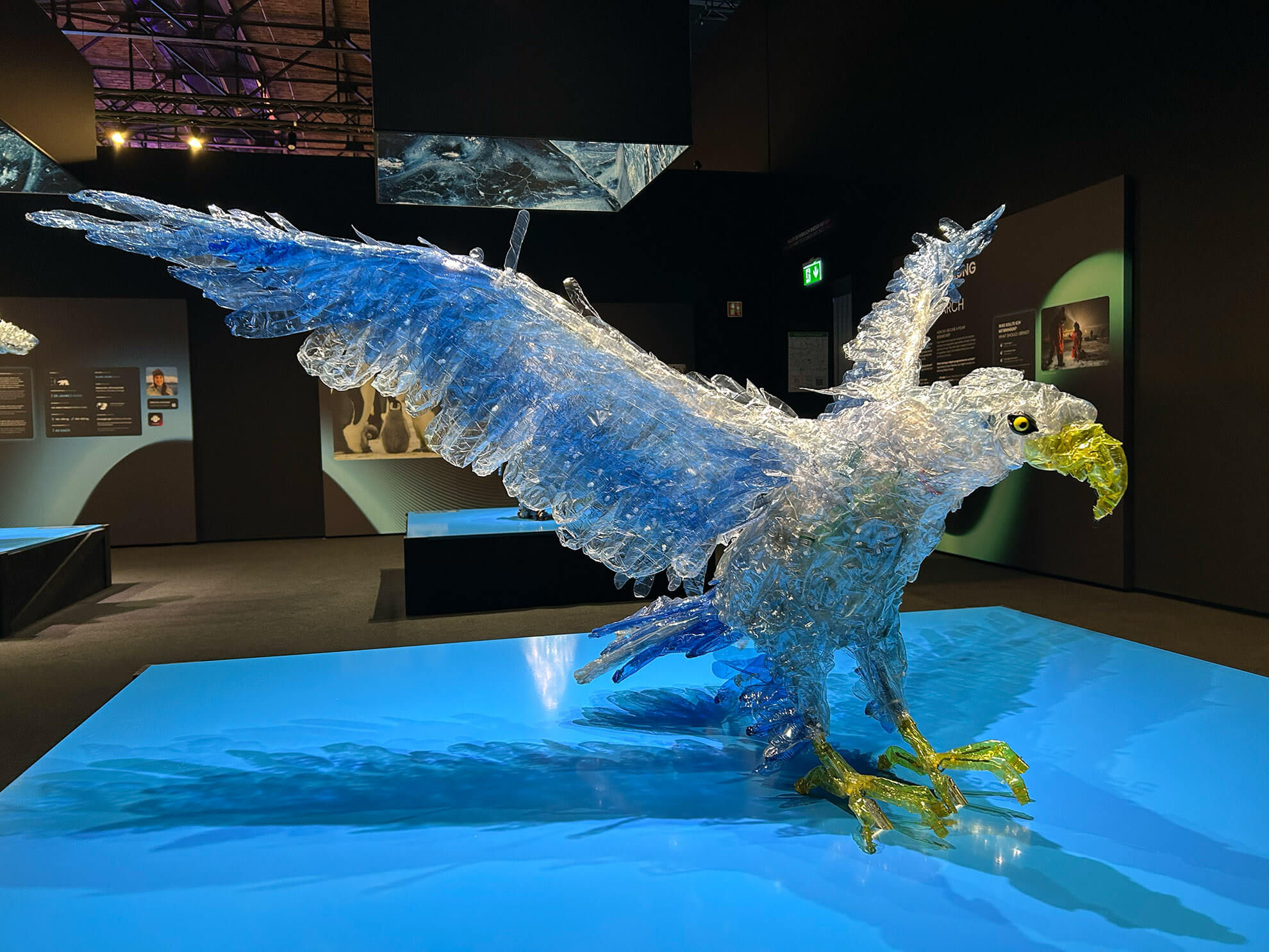 Cosmopolitanschool Berlin Eine große Adlerskulptur mit dem Titel "automatisch gespeicherter Entwurf" ist aus transparenten und blauen Plastikflaschen gefertigt. Er steht auf einem blauen Podest in einem modernen Ausstellungsraum, mit weit ausgebreiteten Flügeln und auffallend gelben Schnabel und Krallen.