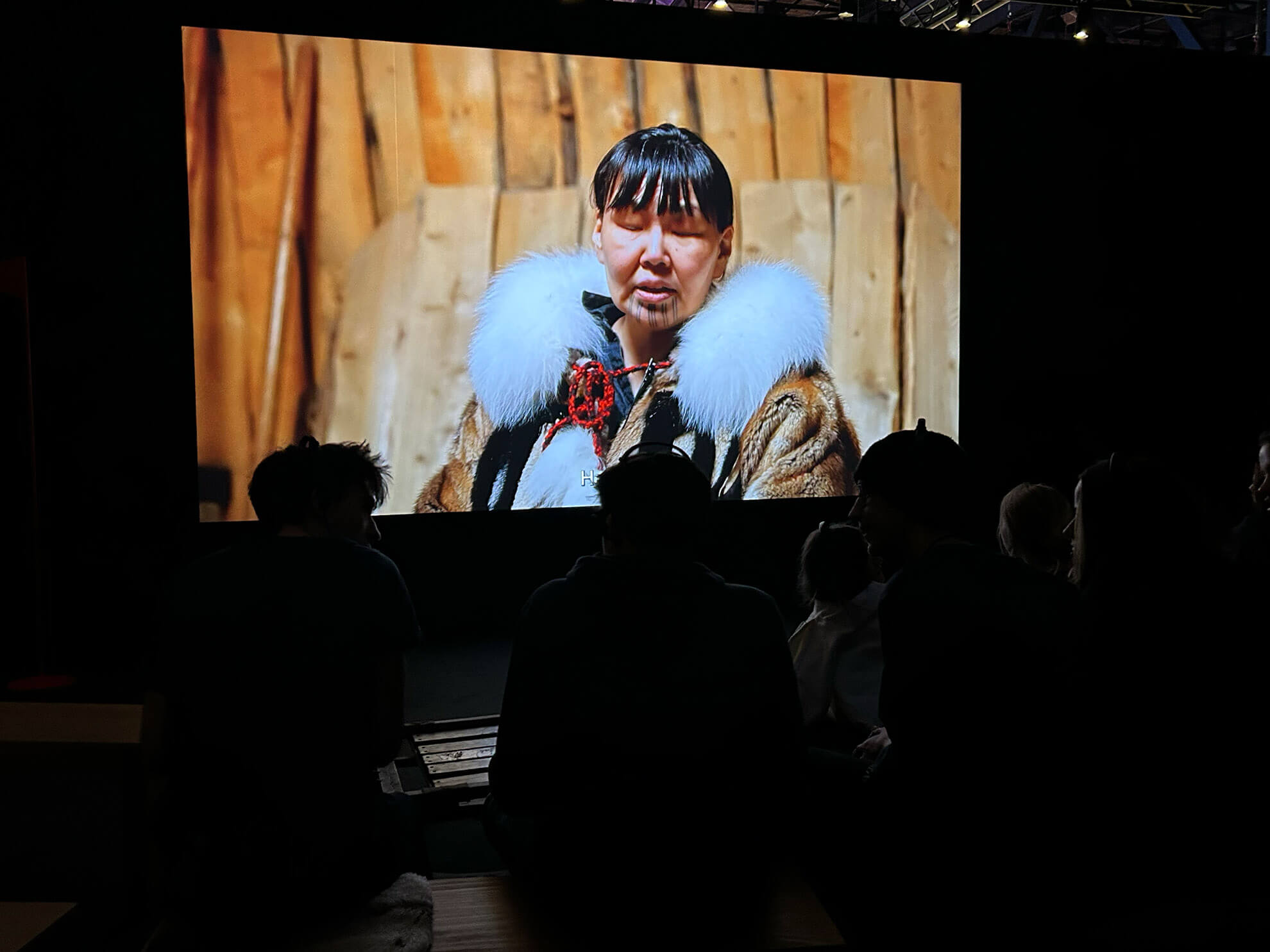 Cosmopolitanschool Berlin Eine Gruppe von Menschen sitzt im Dunkeln und betrachtet einen großen Bildschirm, auf dem eine Person in traditioneller Kleidung mit Pelzbesatz vor einem hölzernen Hintergrund zu sehen ist - ein automatisch gespeicherter Entwurf des kulturellen Erbes, der sich vor ihnen entfaltet.