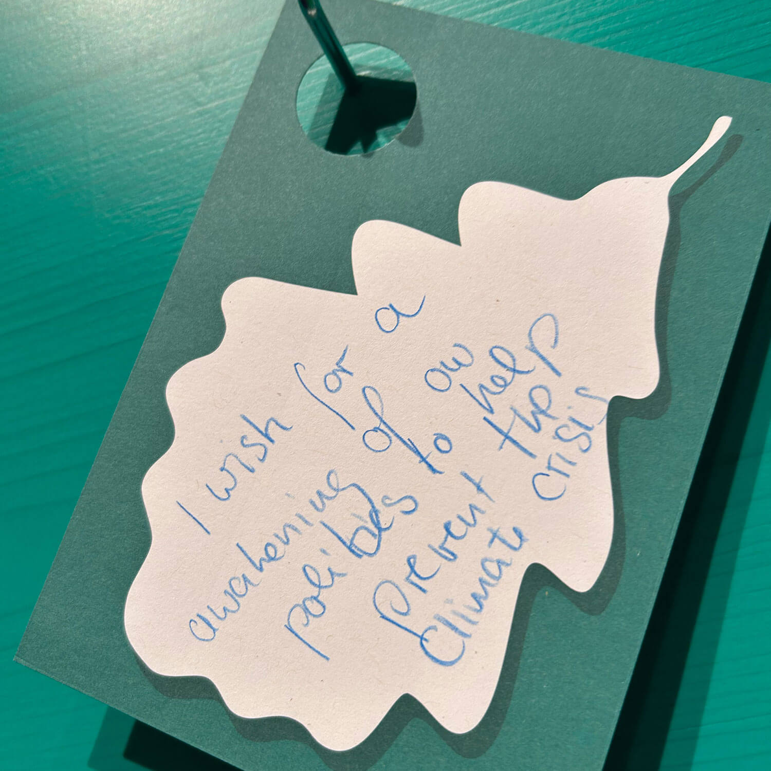 Cosmopolitanschool Berlin Eine blattförmige Papiernotiz mit handgeschriebenem Text, der lautet: Ich wünsche mir ein Aufwachen unserer Politik, um die Klimakrise zu verhindern, platziert auf einer blaugrünen Fläche, die als Automatisch gespeicherter Entwurf gekennzeichnet ist.