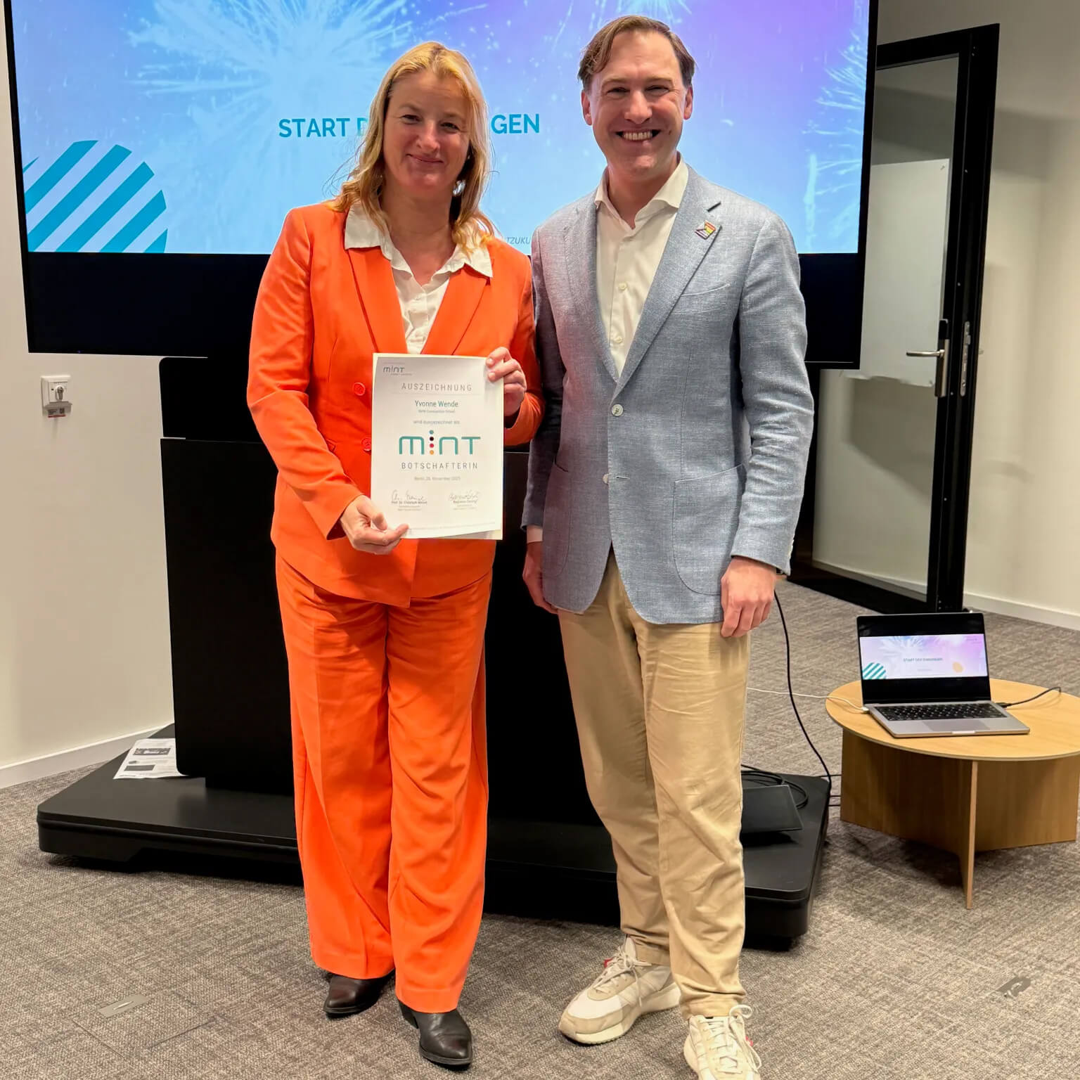 Cosmopolitanschool Berlin Zwei Personen stehen in einem Haus und lächeln in die Kamera. Die Person auf der linken Seite, MINT-Botschafterin Schulgründerin Yvonne Wende, trägt einen orangefarbenen Anzug und hält eine Urkunde in der Hand. Hinter ihnen sind ein Präsentationsbildschirm und ein Laptop zu sehen. loading=