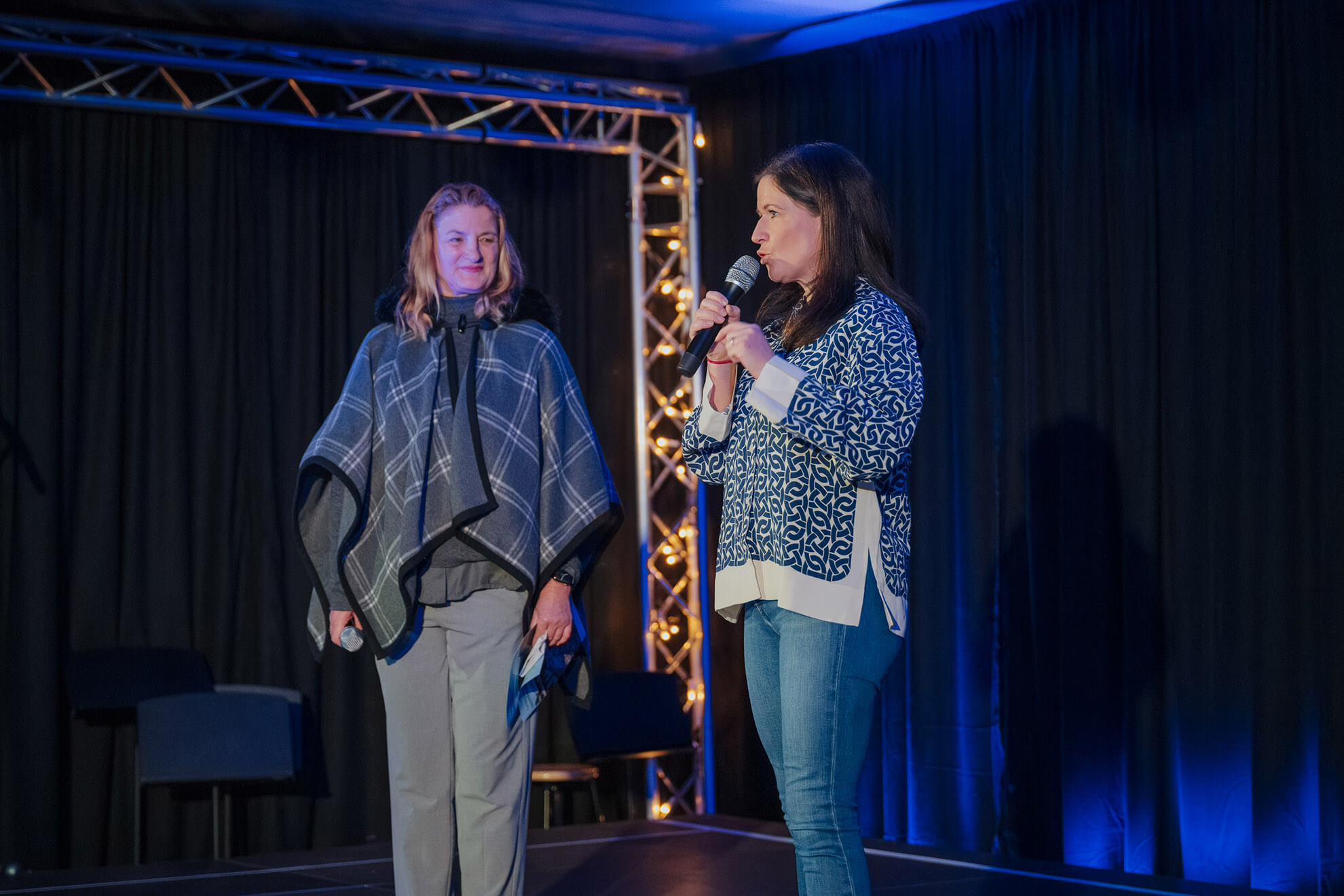 Cosmopolitanschool Berlin Zwei Frauen stehen auf einer Bühne mit dunklen Vorhängen und blauer Beleuchtung. Die eine spricht in ein Mikrofon, die andere hört zu und lächelt, während sie ein gefaltetes Kleidungsstück hält - eine Atmosphäre, die an einen "Automatisch gespeicherten Entwurf" erinnert. Hinter ihnen ist eine beleuchtete Traversenkonstruktion zu sehen. loading=