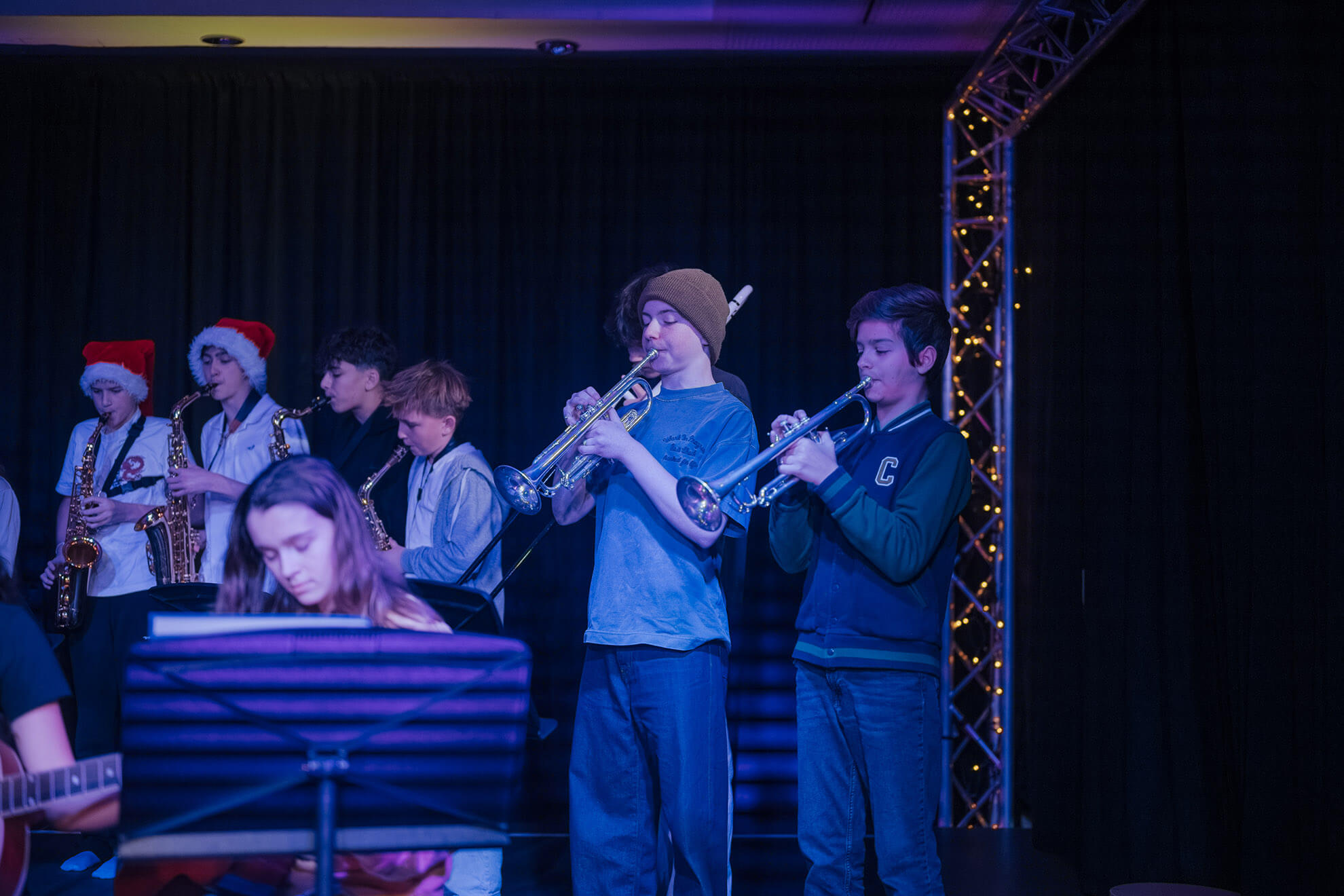 Cosmopolitanschool Berlin Eine Gruppe junger Musiker tritt auf der Bühne auf; zwei Trompeter stehen vorne, während andere im Hintergrund Saxophon spielen - einige mit Weihnachtsmannmützen. Ein Pianist ist teilweise im Vordergrund zu sehen, als wäre er in einem automatisch gespeicherten Entwurf festgehalten.