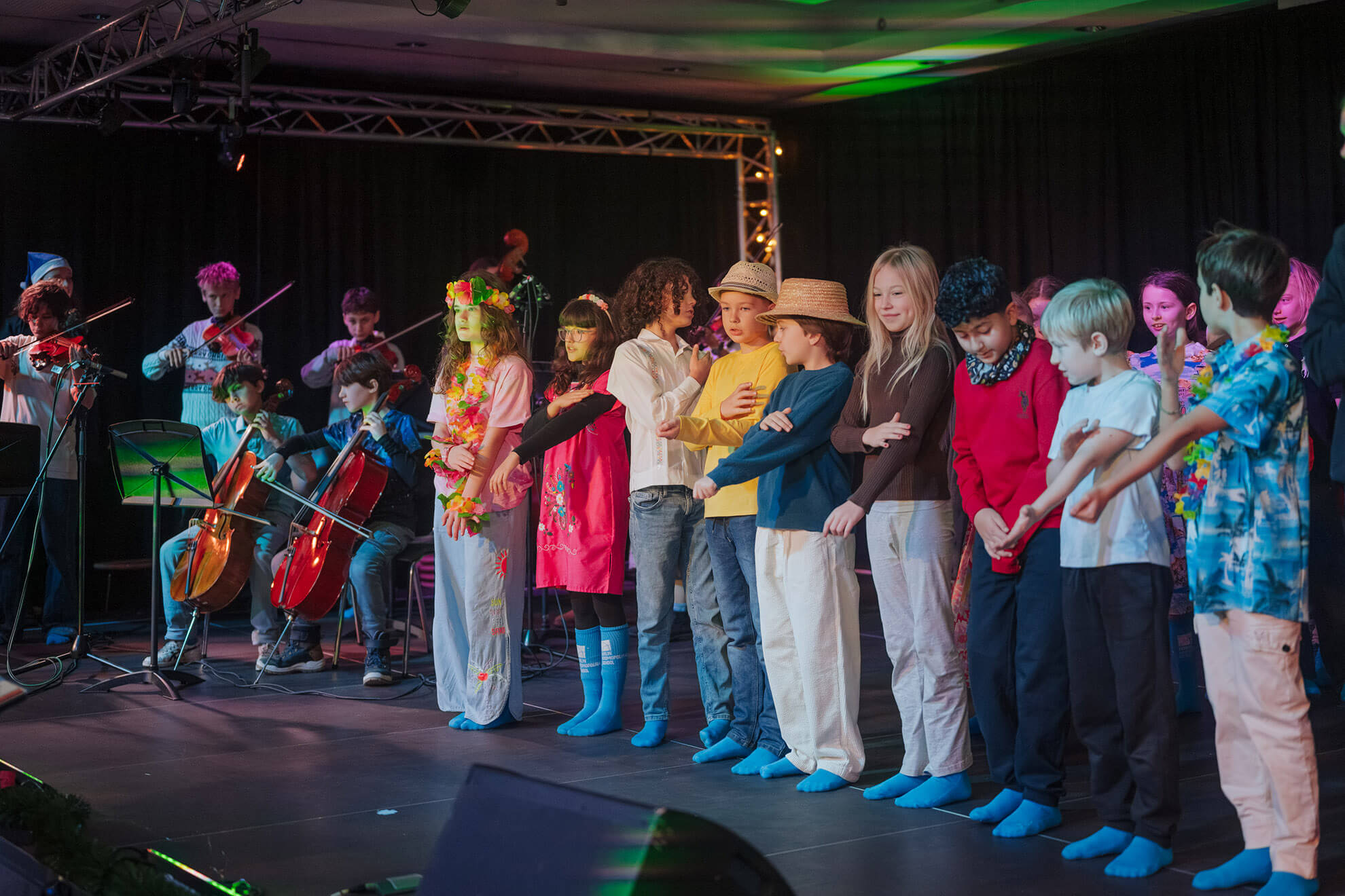 Cosmopolitanschool Berlin Eine Gruppe von Kindern steht auf der Bühne, einige in bunten Kostümen und blauen Socken, während andere im Hintergrund auf Streichinstrumenten spielen. Diese harmonische Szene entfaltet sich im Bühnenlicht - ein automatisch gespeicherter Entwurf jugendlicher Kreativität.