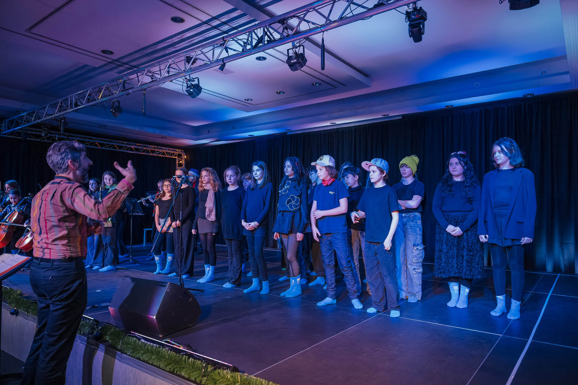 Cosmopolitanschool Berlin Ein Chor von Menschen steht in einer Reihe auf einer Bühne und blickt auf einen Dirigenten, der mit erhobenen Händen gestikuliert. Die Bühne ist mit blauen und violetten Lichtern beleuchtet, und ein schwarzer Vorhang dient als Kulisse.