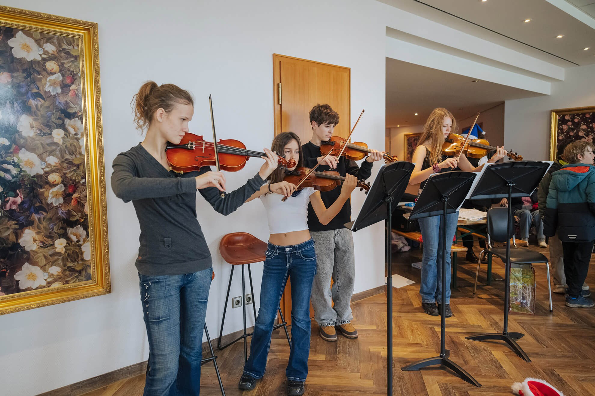 Cosmopolitanschool Berlin Vier junge Leute spielen in einem gut beleuchteten Raum zusammen Geige und stehen neben Notenständern. Zwei große Blumengemälde hängen an den Wänden, und im Hintergrund sind weitere Personen zu sehen.