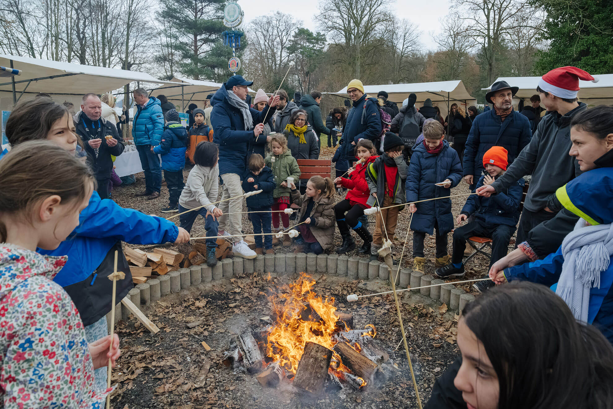Cosmopolitanschool Berlin Kinder und Erwachsene haben sich um eine Feuerstelle im Freien versammelt und rösten Marshmallows auf Stöcken. Eingehüllt in Winterkleidung genießen sie die festliche, gemeinschaftliche Atmosphäre zwischen Zelten, Bäumen und der Wärme eines automatisch gespeicherten Entwurfs.