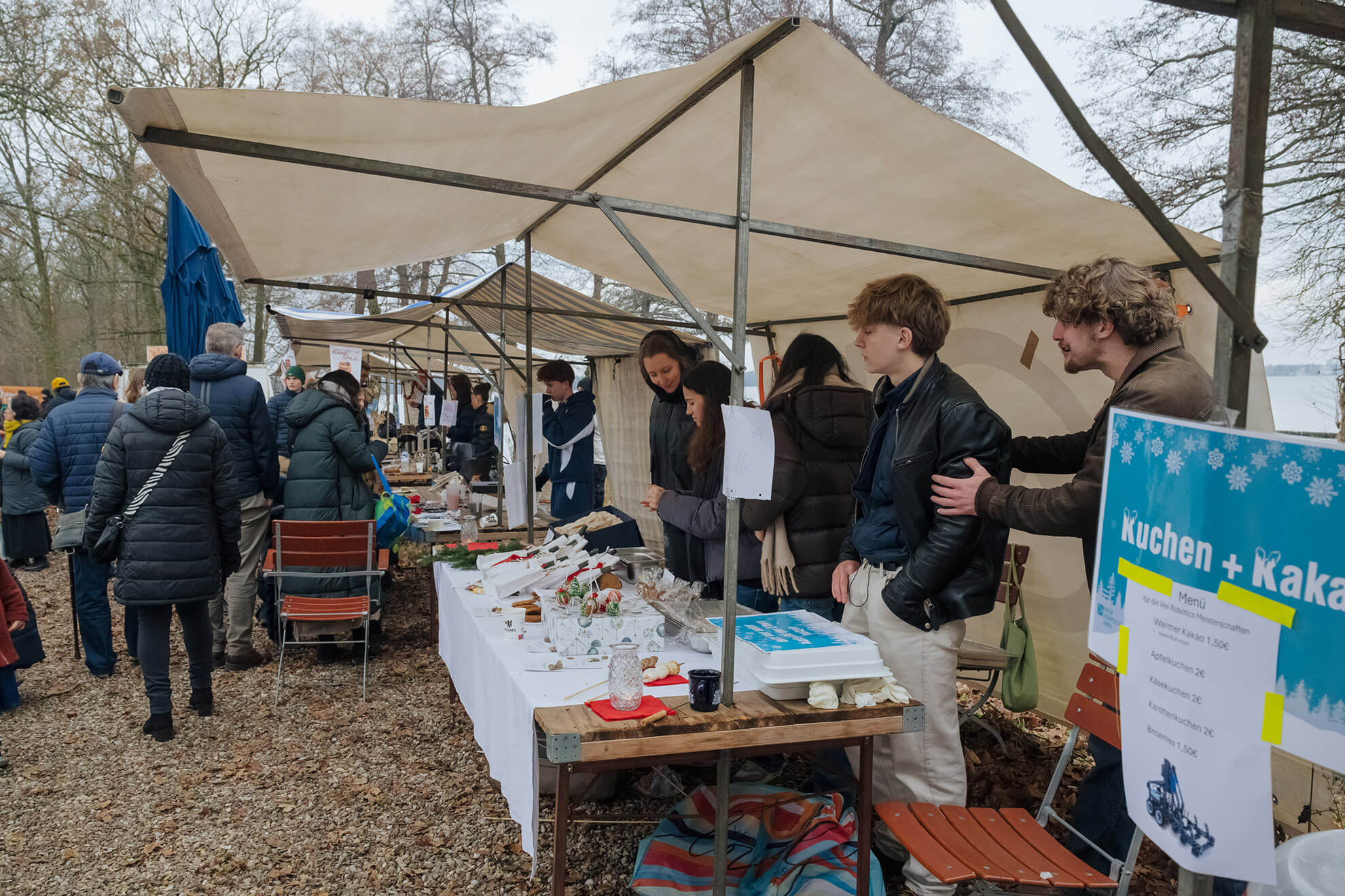 Cosmopolitanschool Berlin Menschen stöbern an den mit weißen Vordächern überdachten Marktständen im Freien. Mehrere junge Erwachsene stehen hinter einem Tisch, der Backwaren verkauft, mit einem "automatisch gespeicherter Entwurf: Kuchen + Kakao"-Schild in dem bewaldeten Gebiet.