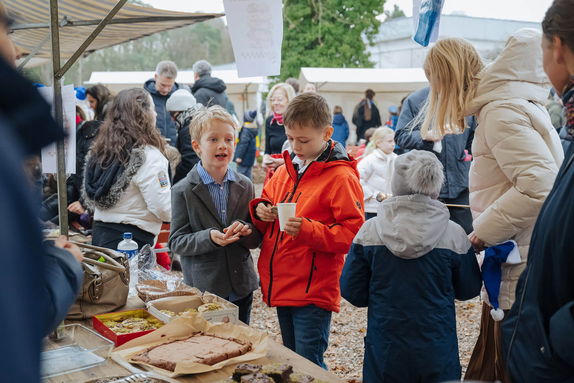 Cosmopolitanschool Berlin Zwei junge Burschen stehen an einem Markttisch mit Backwaren, der eine in einer grauen Jacke, der andere in einem roten Mantel. Um sie herum versammeln sich warm gekleidete Menschen, die einen automatisch gespeicherten Entwurf einer festlichen, geschäftigen Atmosphäre schaffen.