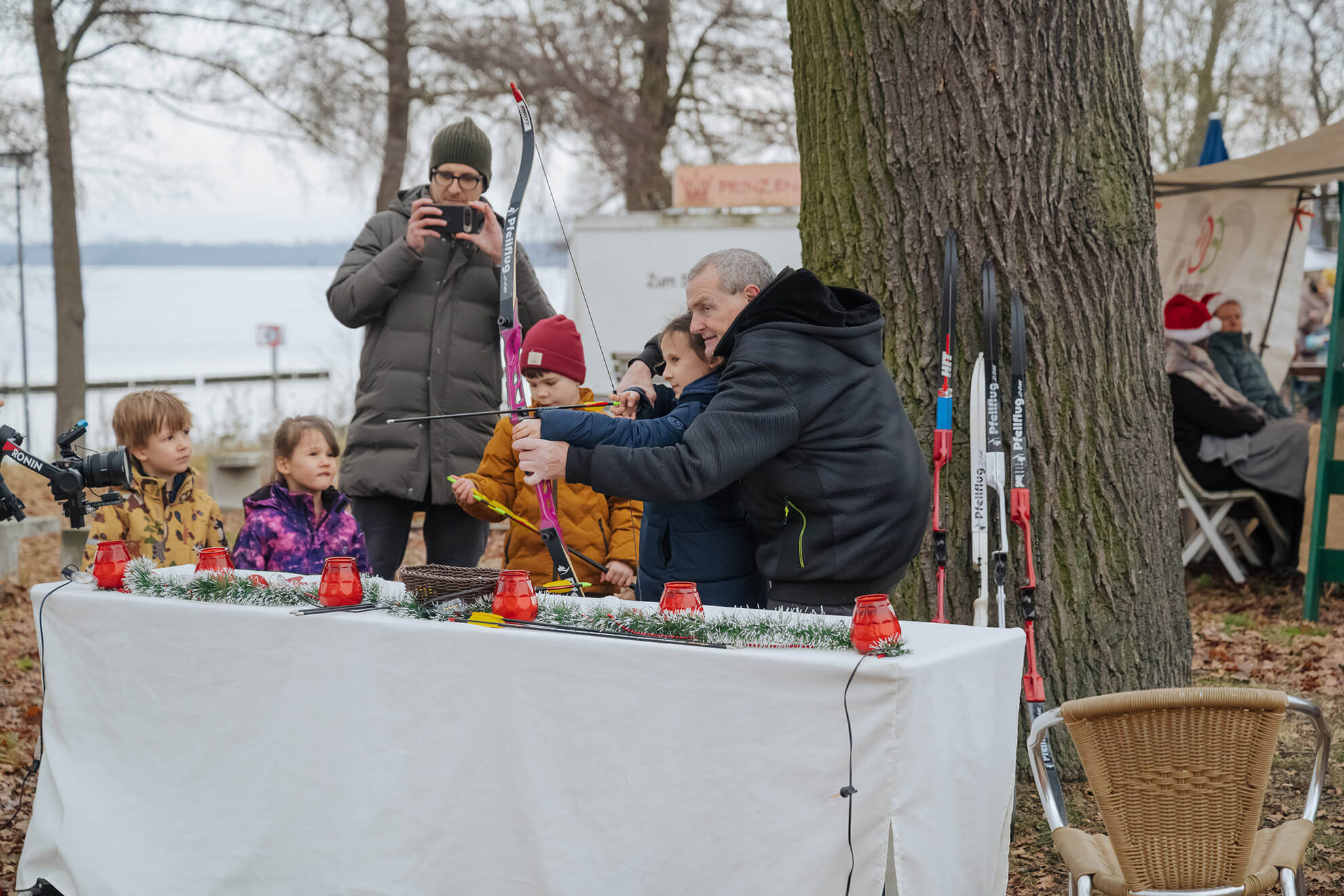 Cosmopolitanschool Berlin Ein Mann hilft einem Kind beim Zielen mit Pfeil und Bogen bei einer Veranstaltung im Freien in der Nähe eines Sees mit Winterbäumen. Automatisch gespeicherter Entwurf: Andere Kinder beobachten, wie festliche Dekorationen einen Tisch im Vordergrund schmücken.
