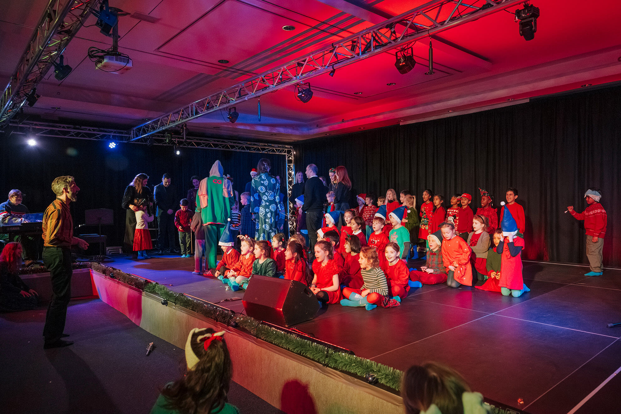 Cosmopolitanschool Berlin Kinder in farbenfrohen Kostümen sitzen und stehen auf einer mit festlichen Lichtern geschmückten Bühne, während Erwachsene und Mitarbeiter, einige in Kostümen, sie organisieren und anleiten. Der Raum ist mit leuchtend roter und blauer Bühnenbeleuchtung beleuchtet. loading=
