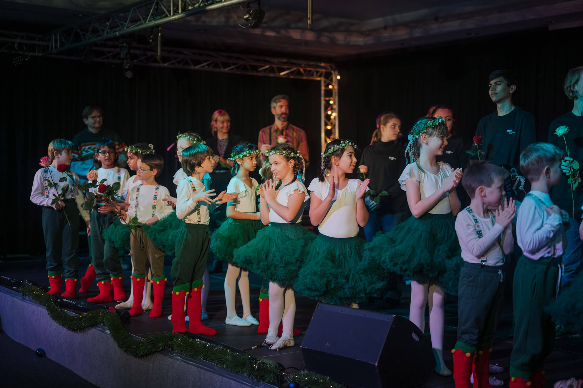 Cosmopolitanschool Berlin Eine Gruppe von Kindern in grünen Tutus, weißen Oberteilen und Blumenkränzen und Jungen in weißen Hemden, grünen Hosen und roten Stiefeln stehen auf der Bühne, halten Blumen und klatschen, während die Erwachsenen hinter ihnen stehen. loading=